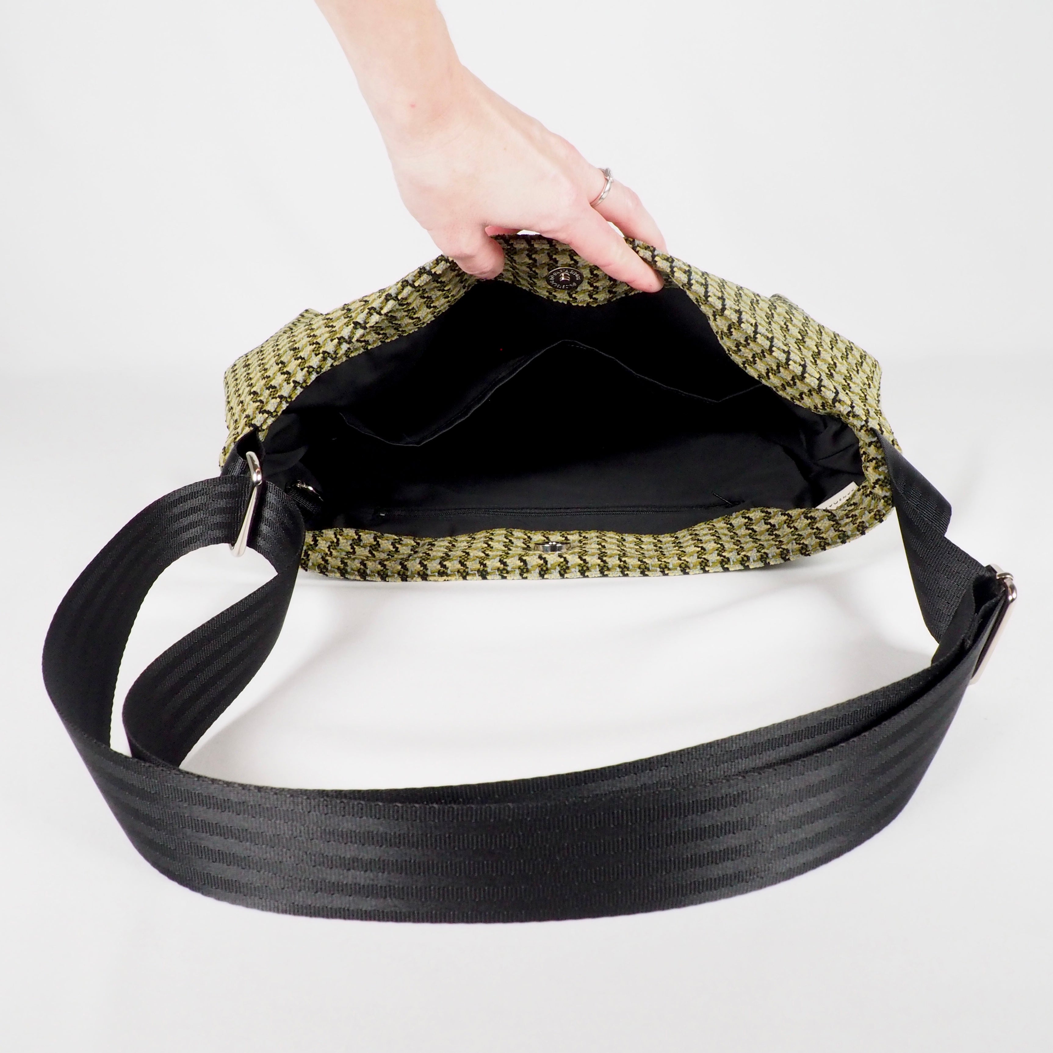 Eine Hand öffnet eine gewebte Tasche mit schwarzen Innenfächern und einem breiten, schwarzen Schulterriemen.