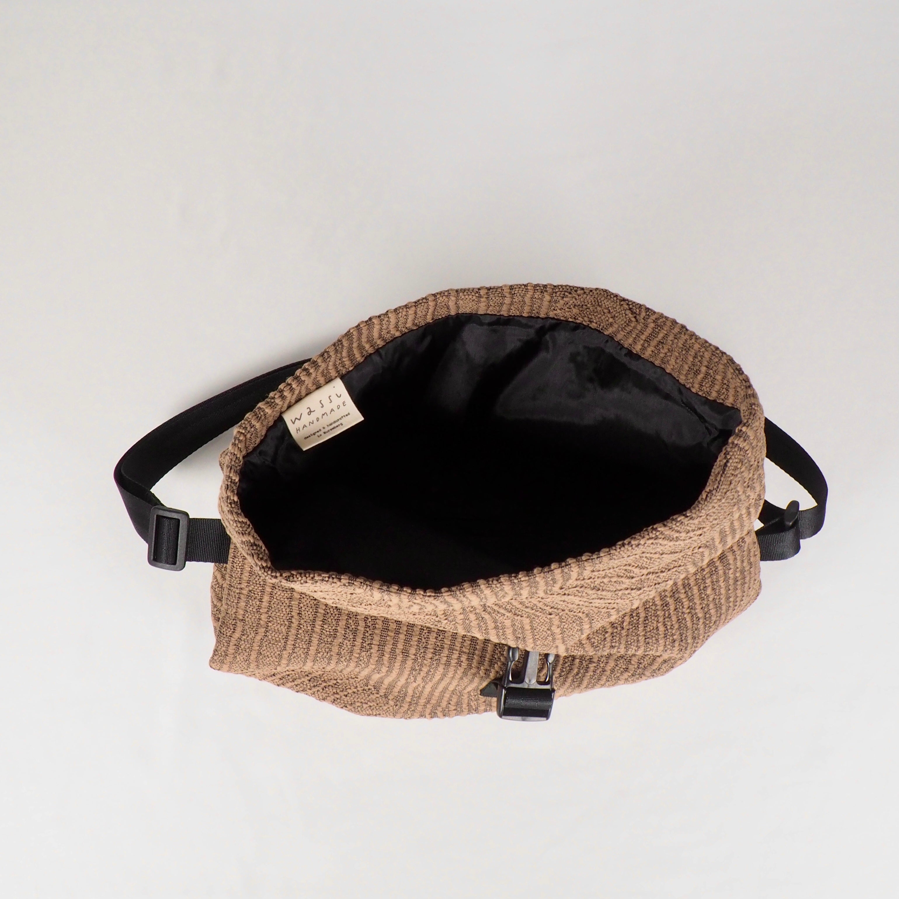 Braune Gürteltasche aus gestricktem Stoff mit schwarzem Innenfutter und einem Etikett im Inneren, auf weißem Hintergrund.