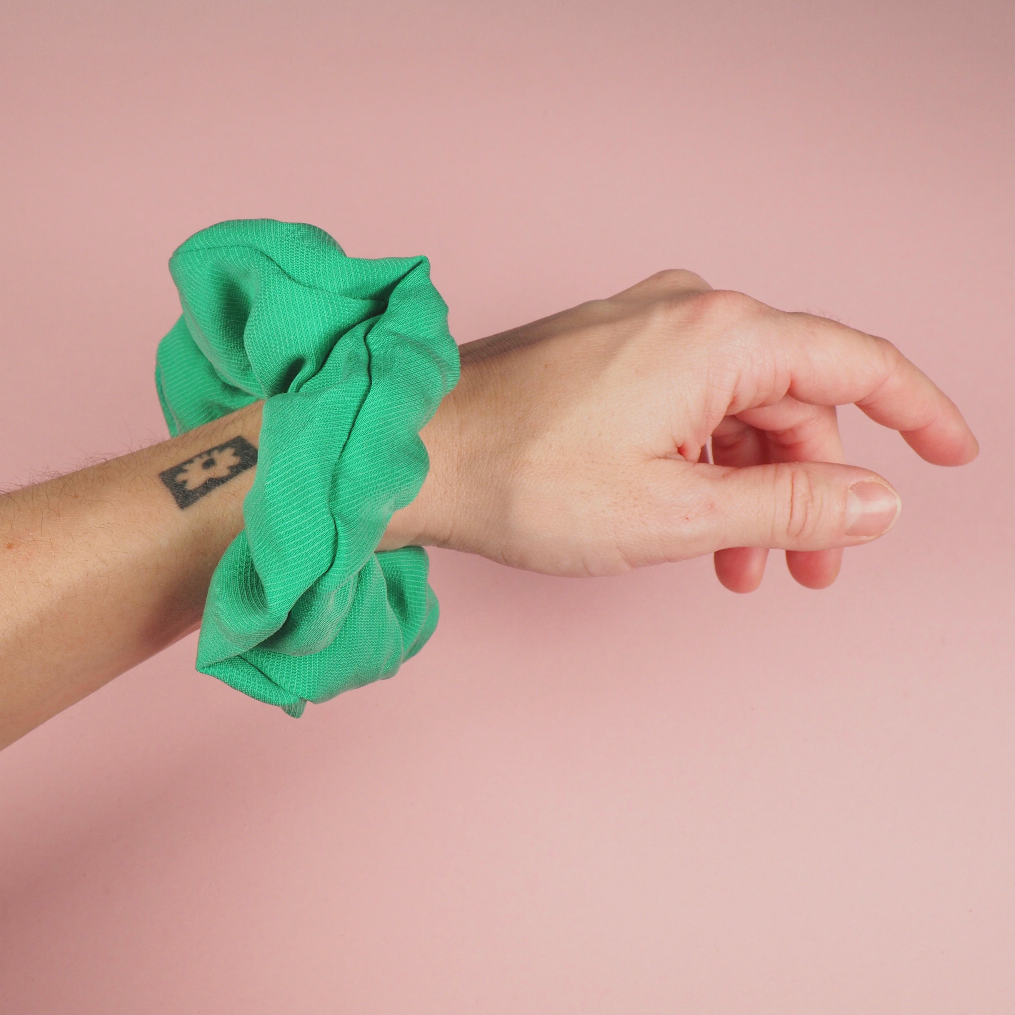 Eine Hand mit einem grünen Scrunchie um das Handgelenk und einem kleinen schwarzen Tattoo, vor einem rosa Hintergrund.