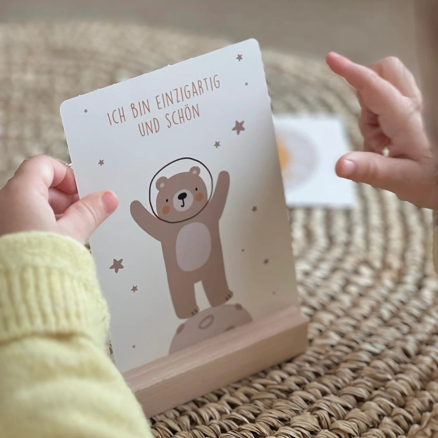 Die Hände eines Kindes zeigen auf eine Karte auf einer gewebten Matte. Die Karte zeigt einen Cartoon-Bären in einem Raumanzug, der auf einem Felsen steht, mit dem deutschen Text "ICH BIN EINZIGARTIG UND SCHÖN.".