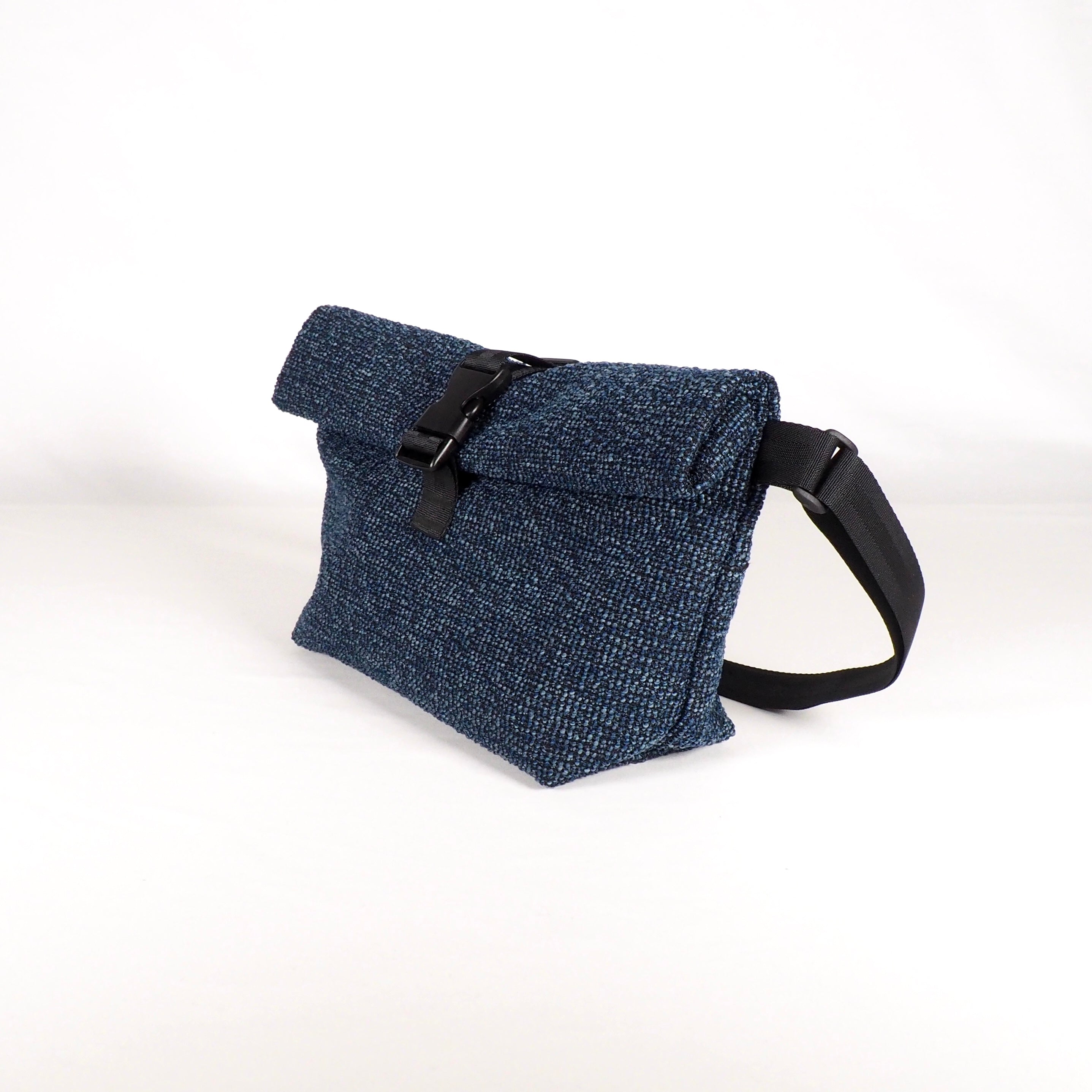 Eine blaue Crossbody-Tasche aus strukturiertem Stoff mit einem umklappbaren Verschluss, der mit einer schwarzen Schnalle und einem verstellbaren schwarzen Riemen gesichert ist, vor einem schlichten weißen Hintergrund.