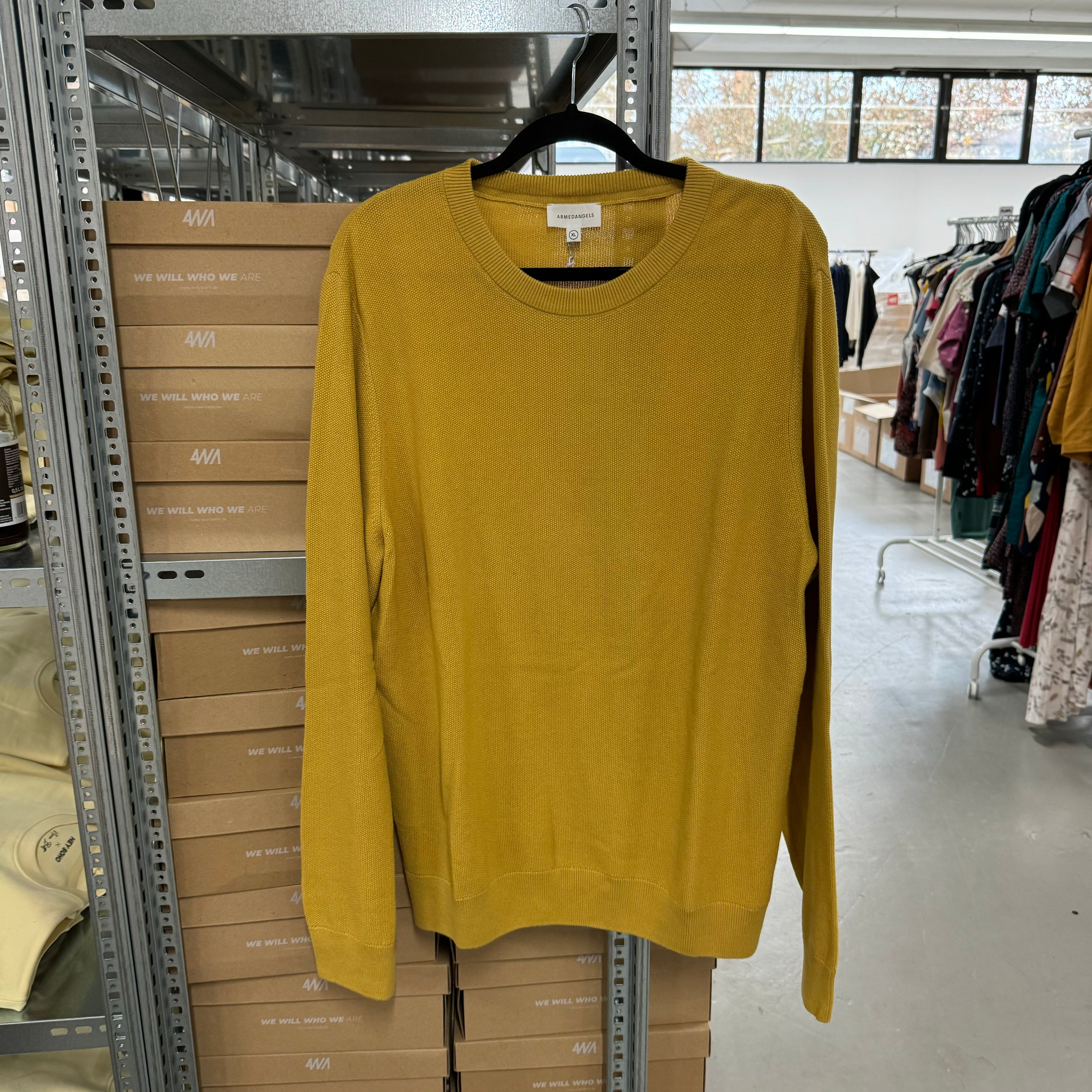 Gelber Pullover auf einem Bügel in einem Lagerraum mit gestapelten Kartons und Kleidern im Hintergrund.