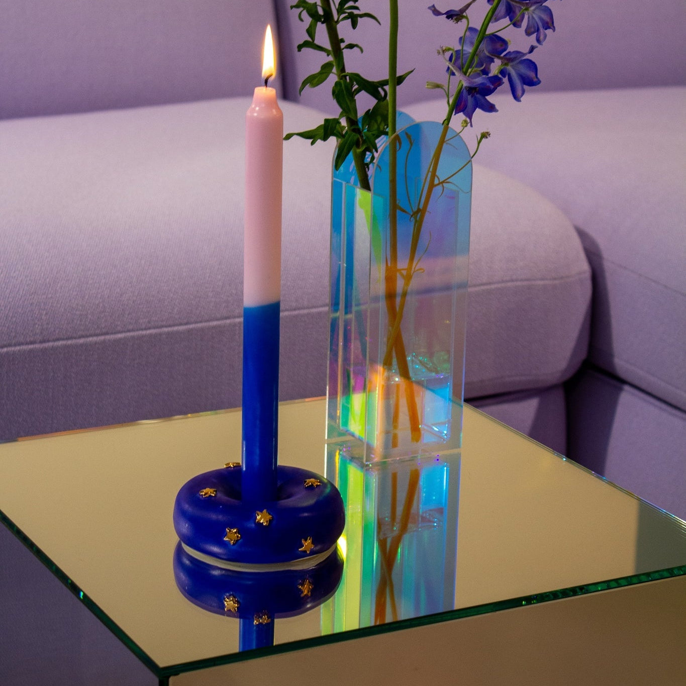 Zweifarbige Kerze in blauem Sternenhalter und Vase mit Blumen auf einem Glastisch vor einem lila Sofa.