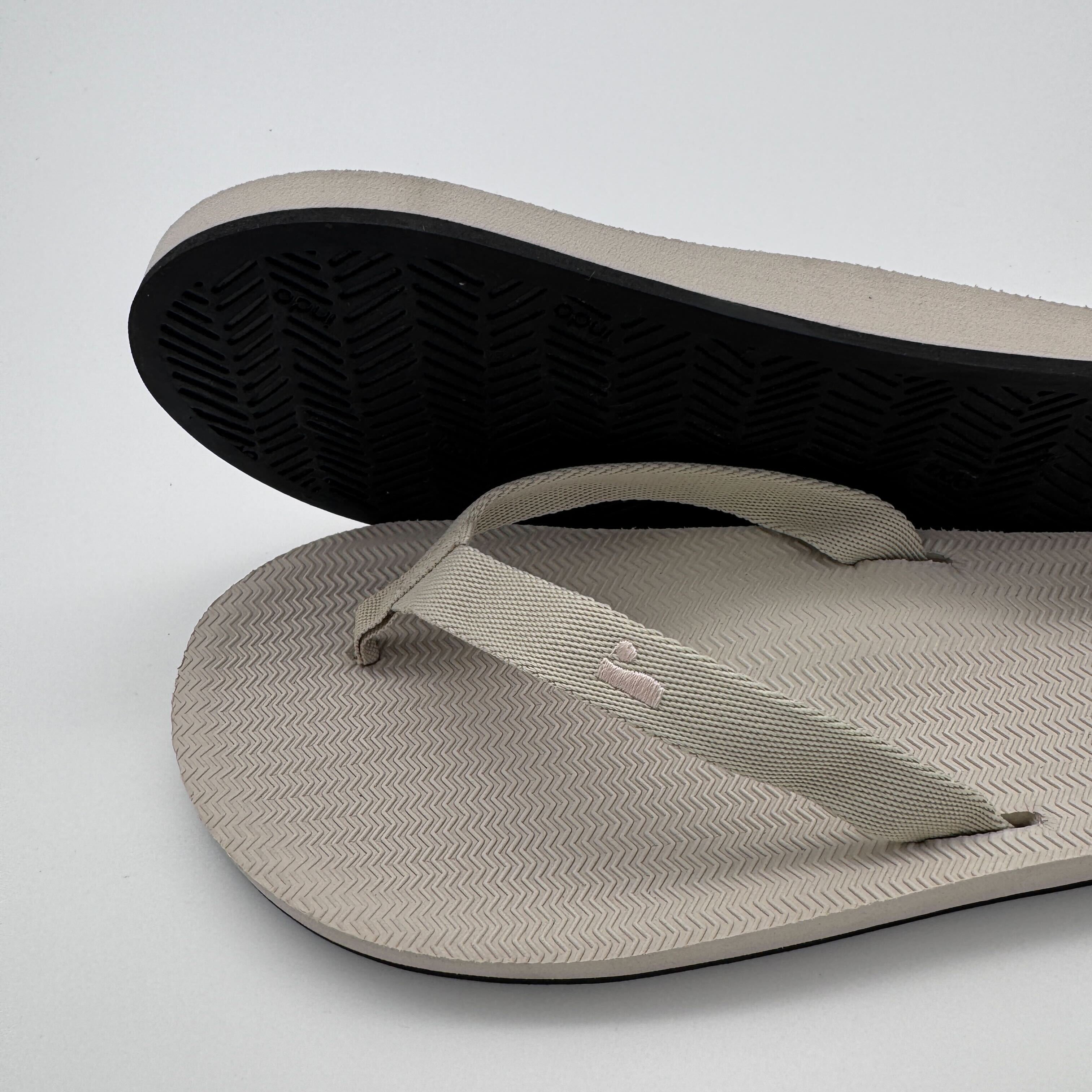 Beige Flip-Flops mit schwarzen Sohlen, übereinander gelegt auf weißem Hintergrund.