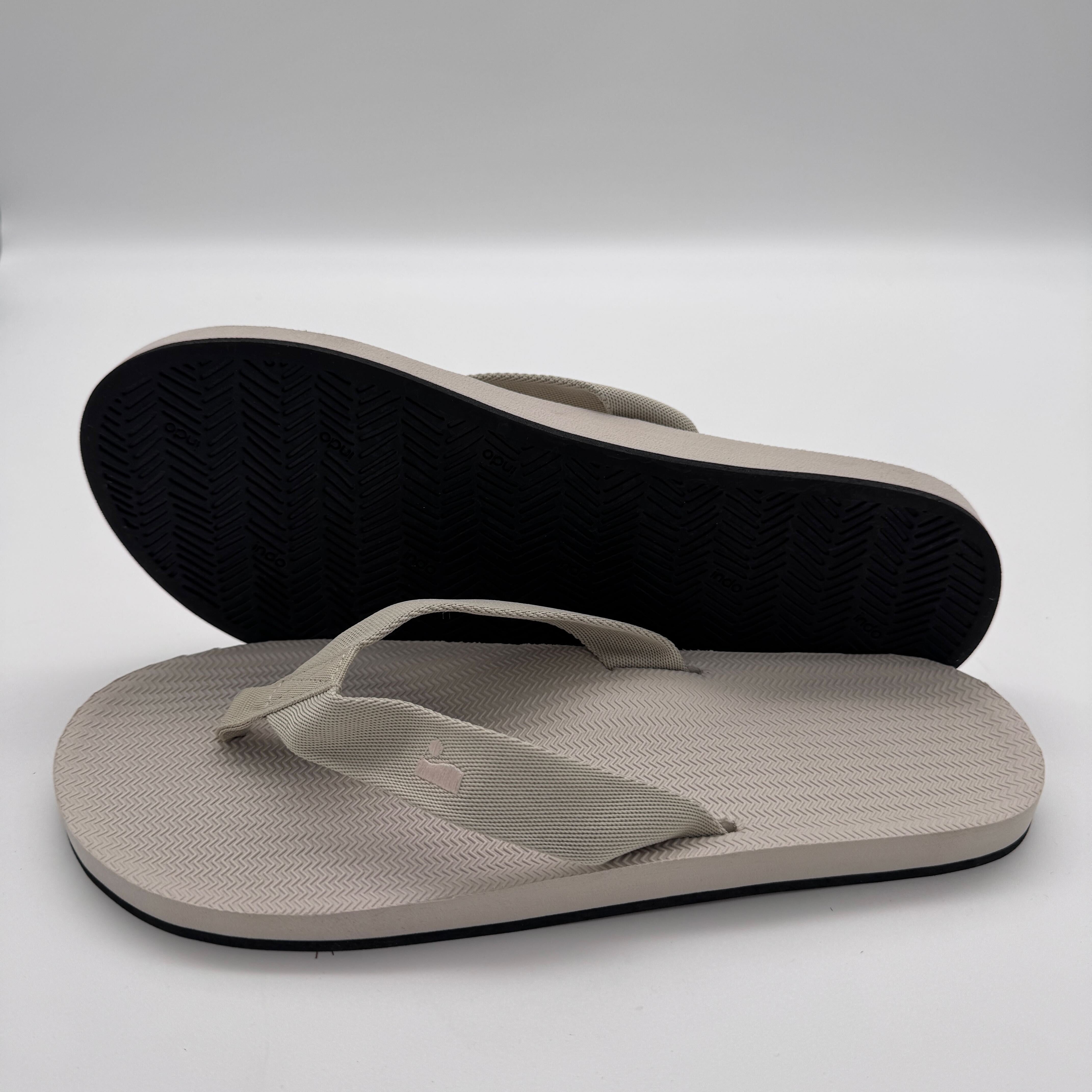 Beige Flip-Flops mit schwarzer Sohle, ein Paar, einer mit der Sohle nach oben liegend, auf weißem Hintergrund.
