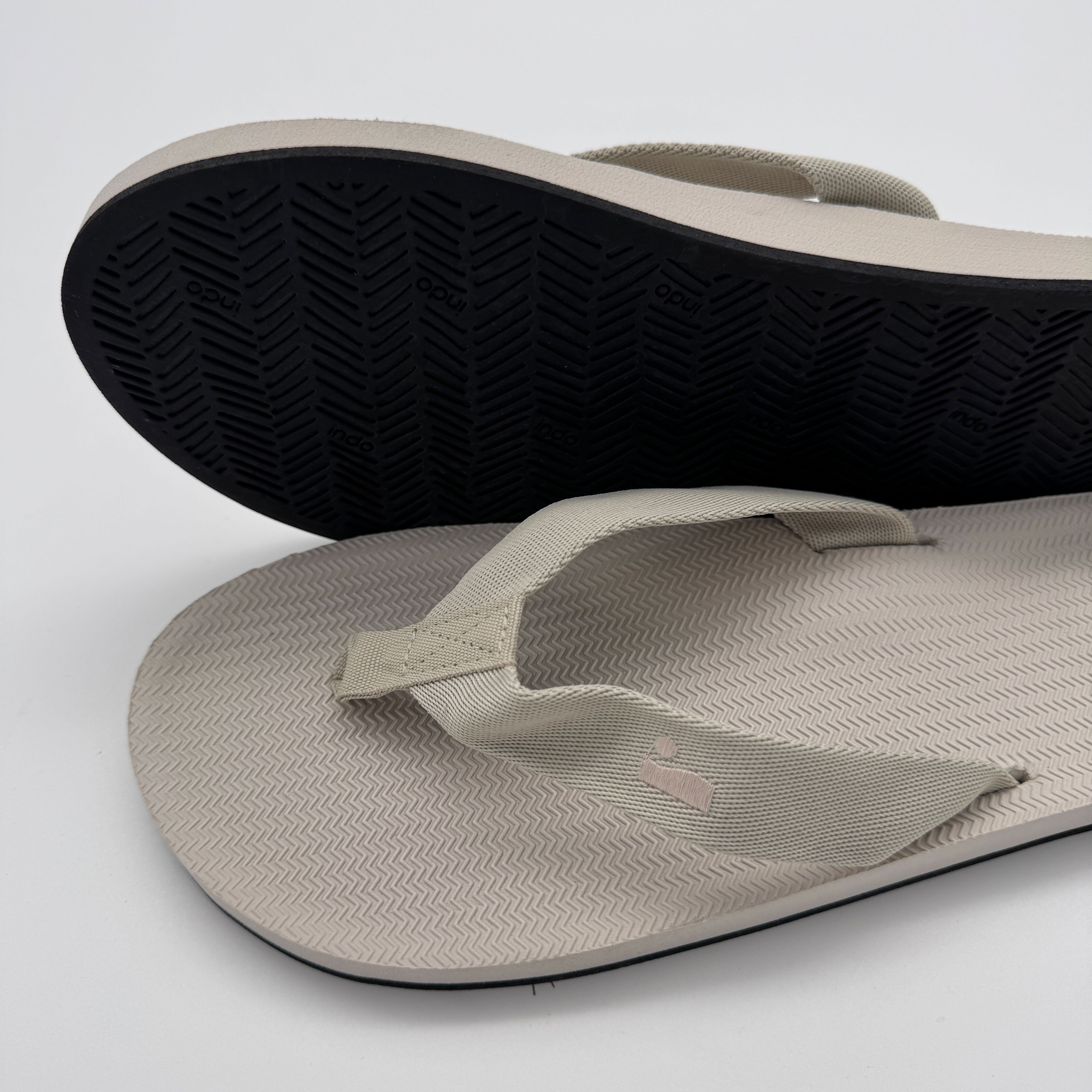Beige Flip-Flops mit schwarzer Sohle, eine umgedreht, auf weißem Hintergrund.