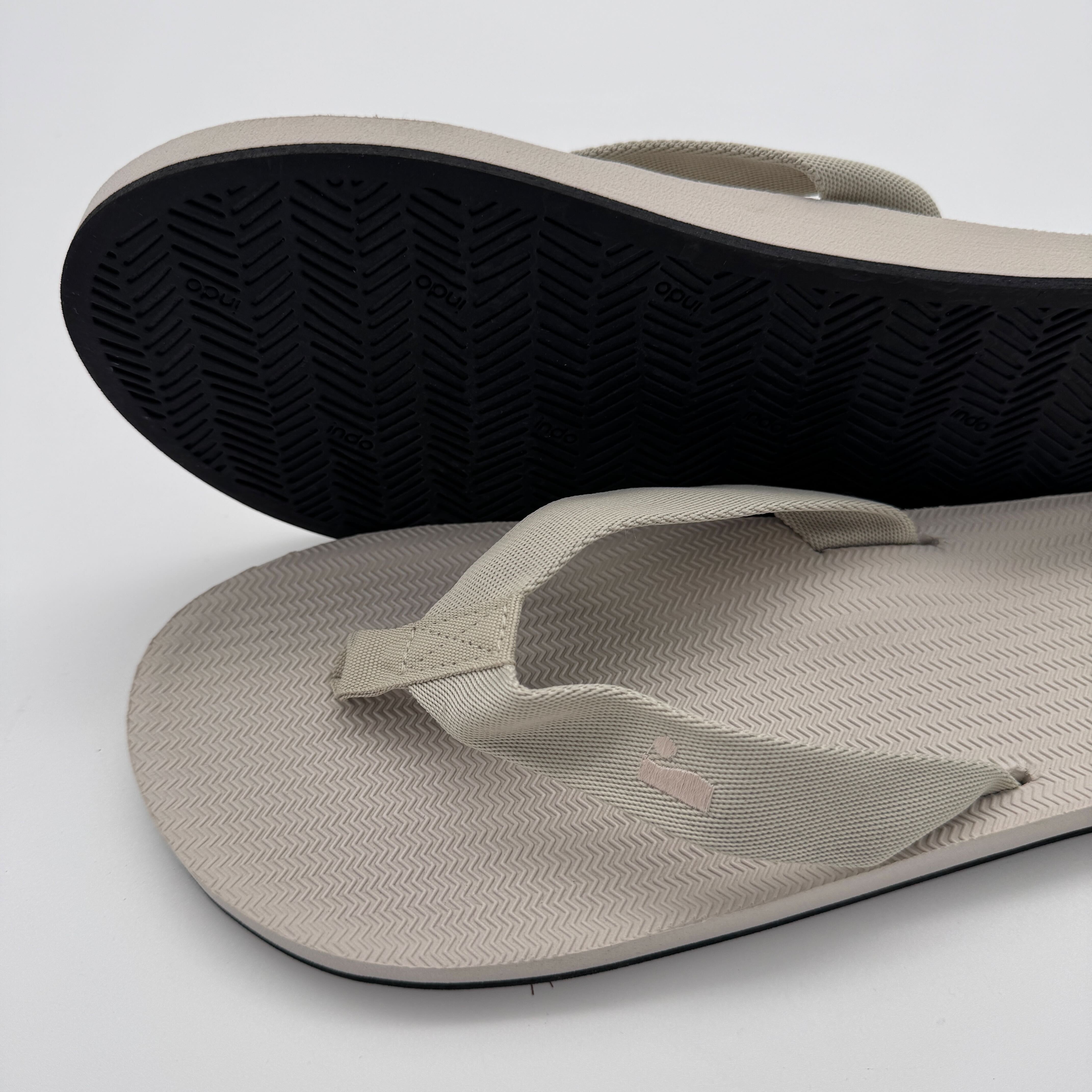 Beige Flip-Flops mit schwarzer Sohle auf weißem Hintergrund, die zur Seite geneigt sind.