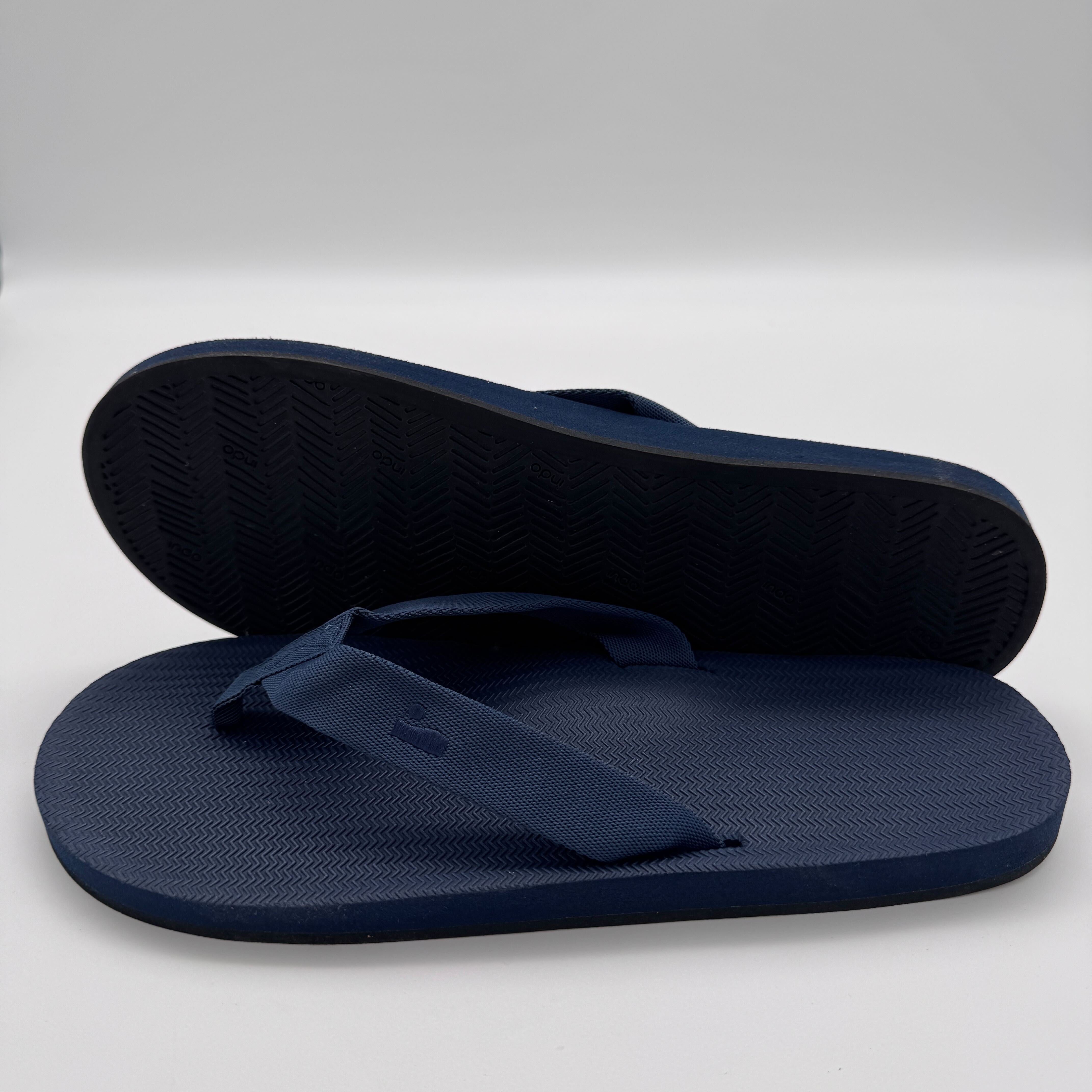 Zwei dunkelblaue Flip-Flops, einer liegt flach, der andere steht aufrecht, auf einem weißen Hintergrund.