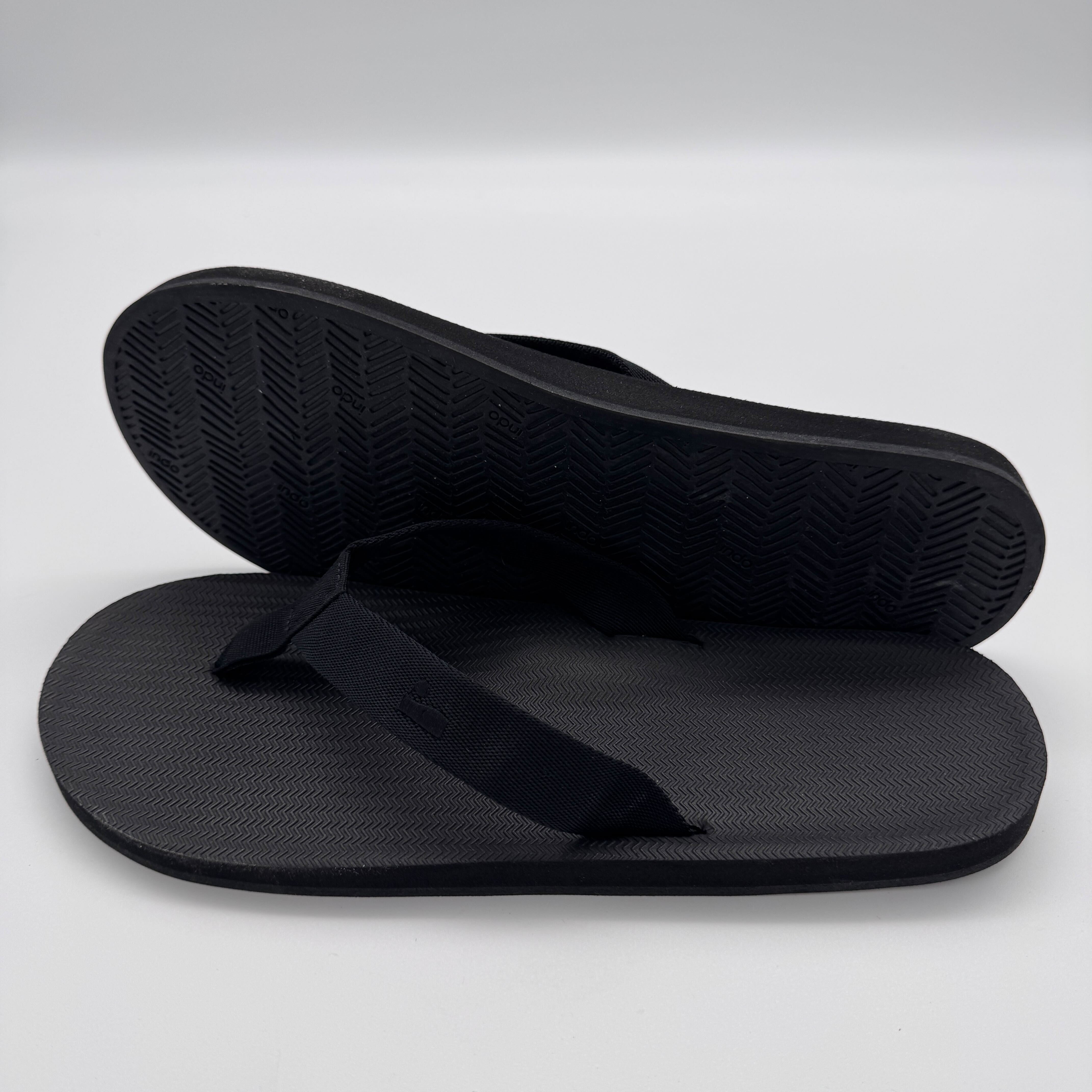 Zwei schwarze Flip-Flops, einer liegt auf einem grauen Hintergrund, der andere zeigt die Sohle mit Fischgrätenmuster.