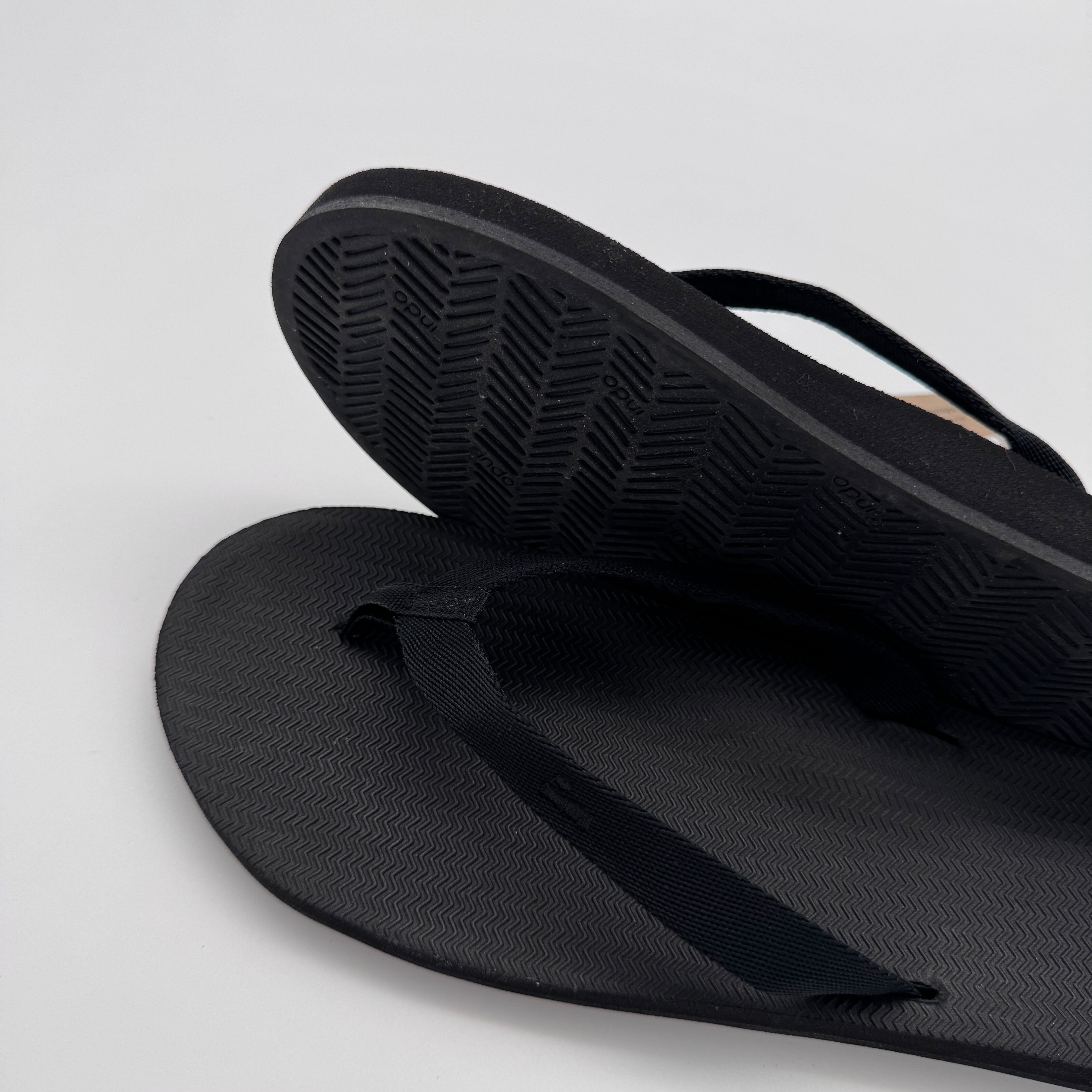 Schwarze Flip-Flops, eine aufrecht mit sichtbarer Sohle, die andere liegend, auf einem neutralen Hintergrund.