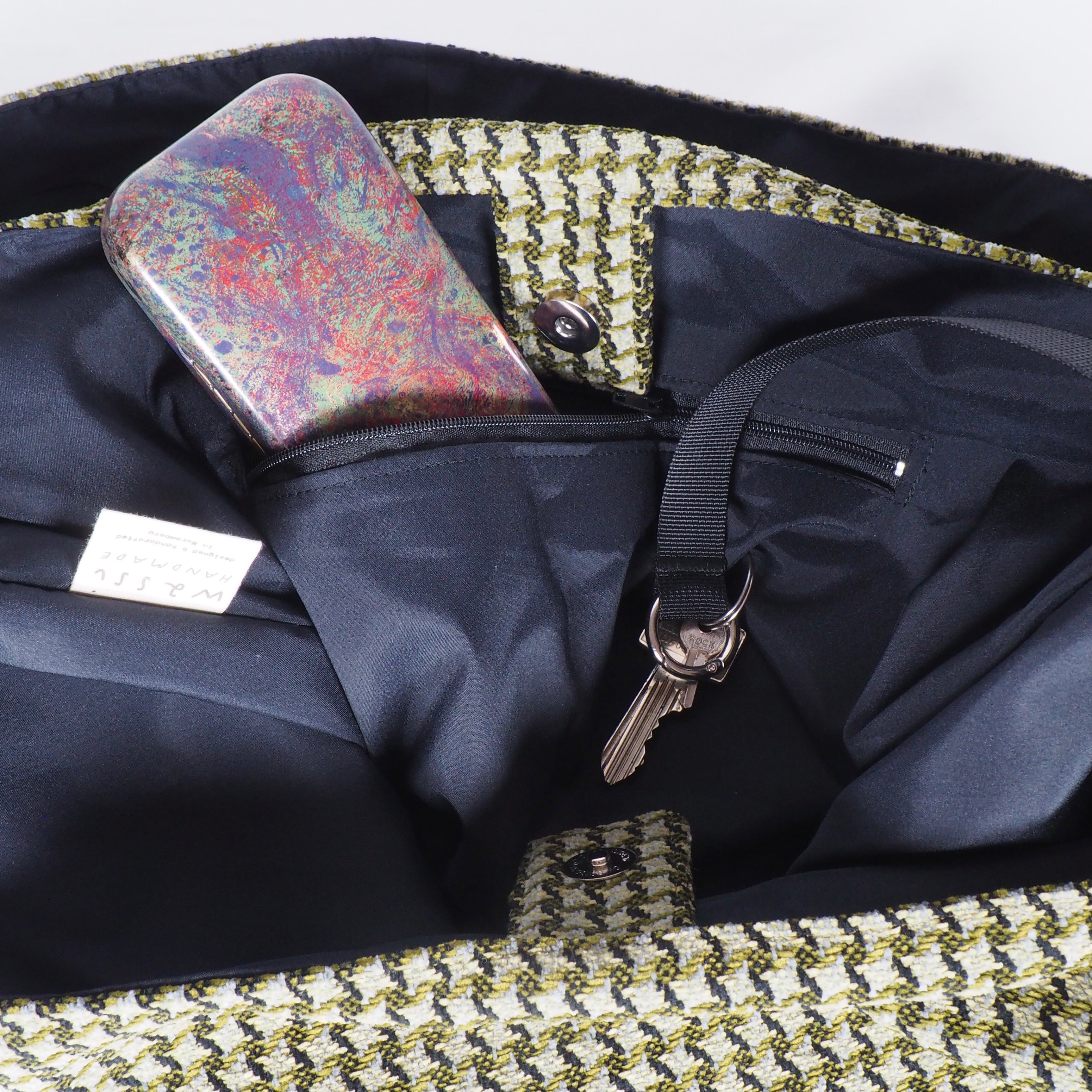 Eine geöffnete Handtasche zeigt ein buntes Telefon in einer Innentasche und einen Schlüsselbund, der an einem schwarzen Riemen befestigt ist, wobei ein schwarzes Stofffutter und eine gemusterte Außenkante sichtbar sind.