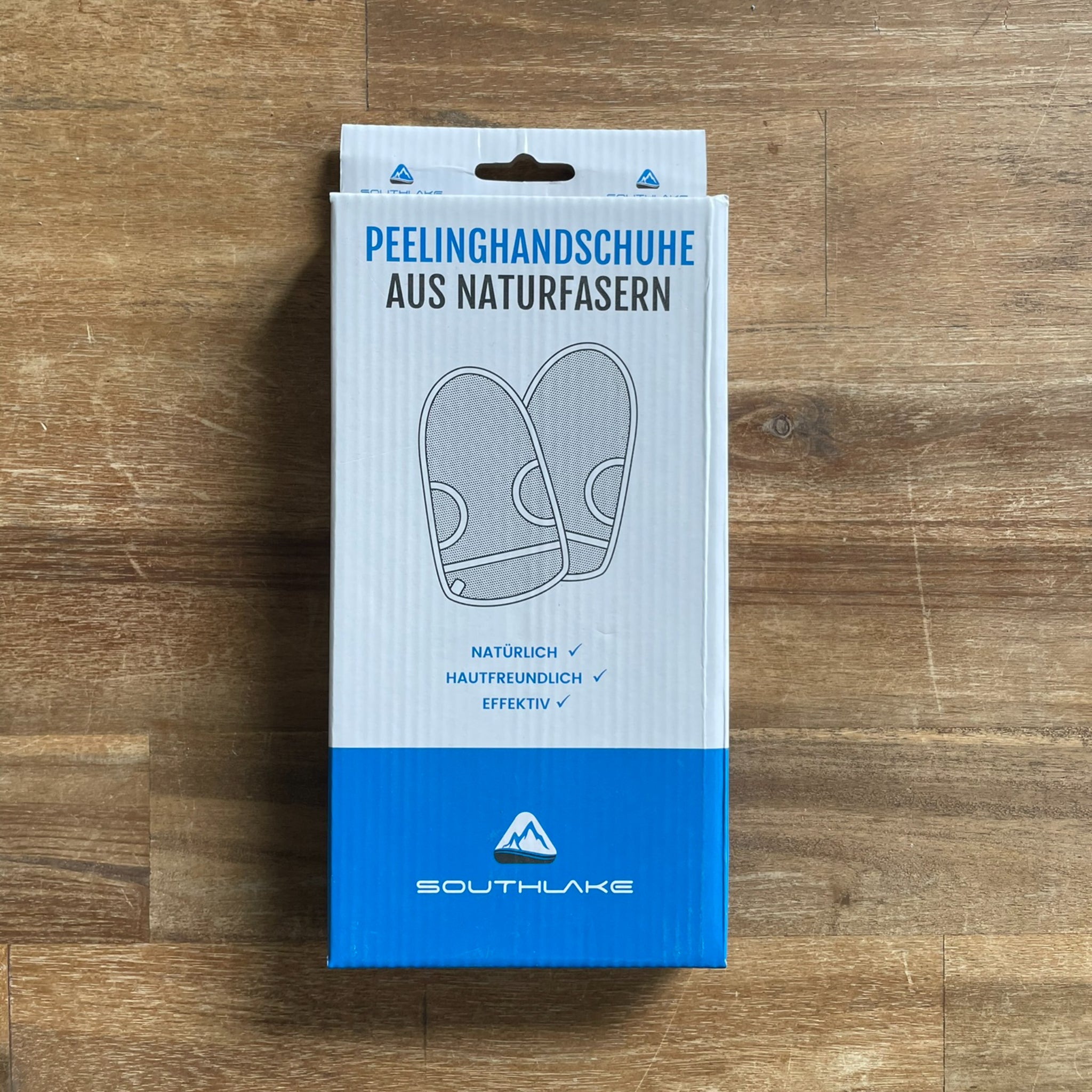 Kartonverpackung von Peelinghandschuhen aus Naturfasern auf einem Holztisch, mit blauer und weißer Aufschrift.