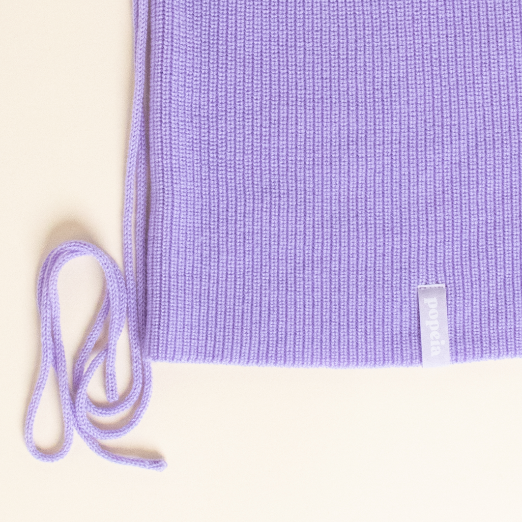 Lila gestricktes Kleidungsstück mit Schnüren und Etikett auf einem cremefarbenen Hintergrund.