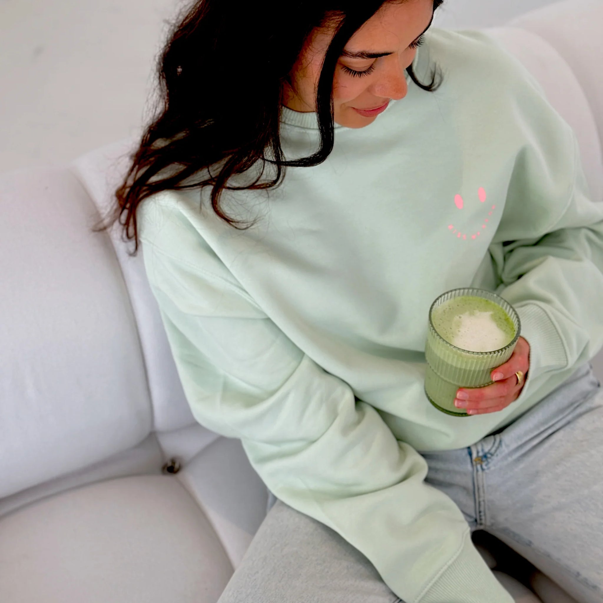 Eine Frau in einem hellgrünen Sweatshirt und Jeans sitzt auf einer weißen Couch, hält ein Glas mit einem grünen Smoothie in der Hand und schaut mit einem leichten Lächeln nach unten.