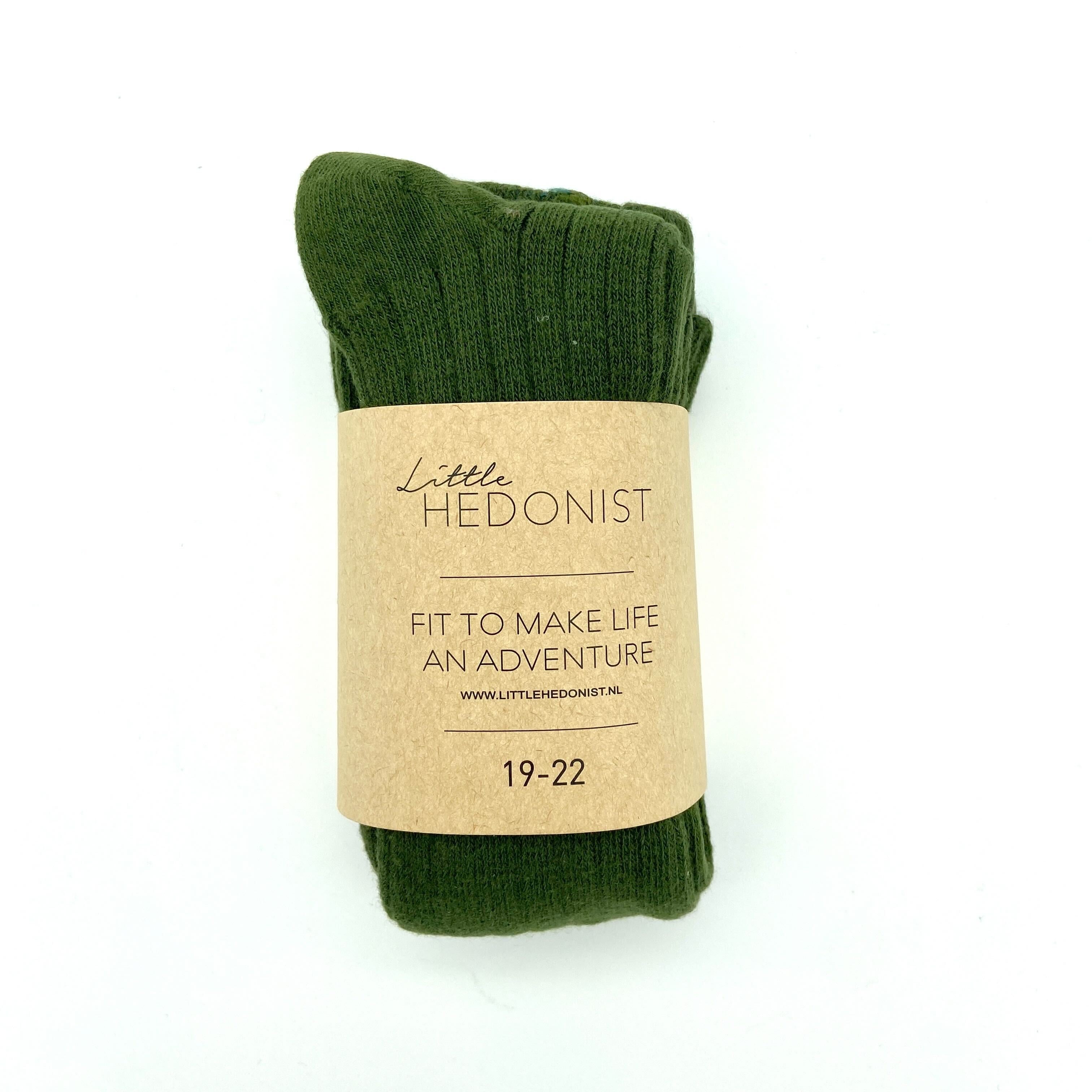 Ein Paar dunkelgrüne Socken mit einem braunen Papieretikett, auf dem "Little Hedonist" steht. Fit, um das Leben zu einem Abenteuer zu machen. 19-22." Die Socken liegen flach auf einem weißen Hintergrund.