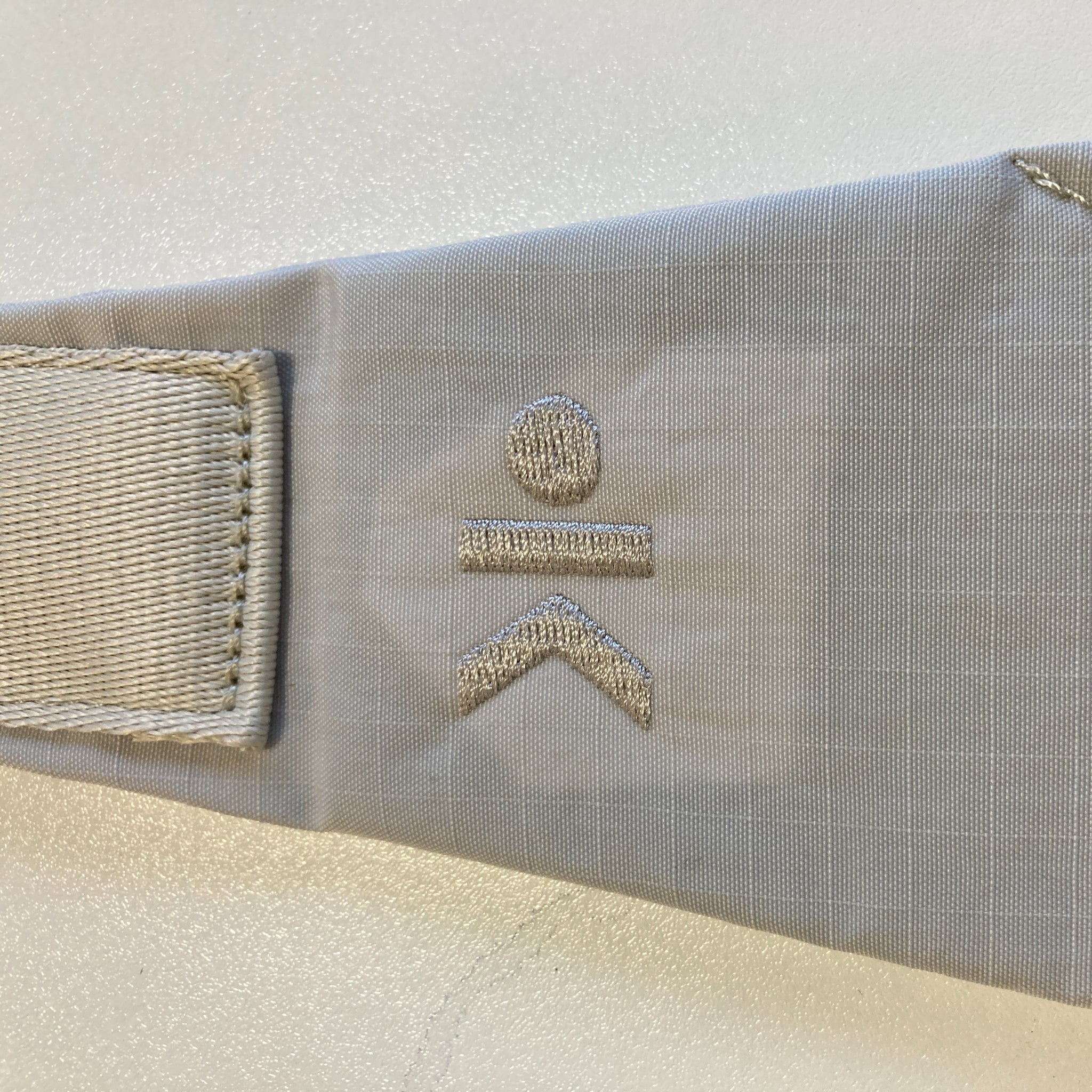 Graues Stoffstück mit gesticktem Symbol: Punkt über einer Linie und einem umgekehrten V.