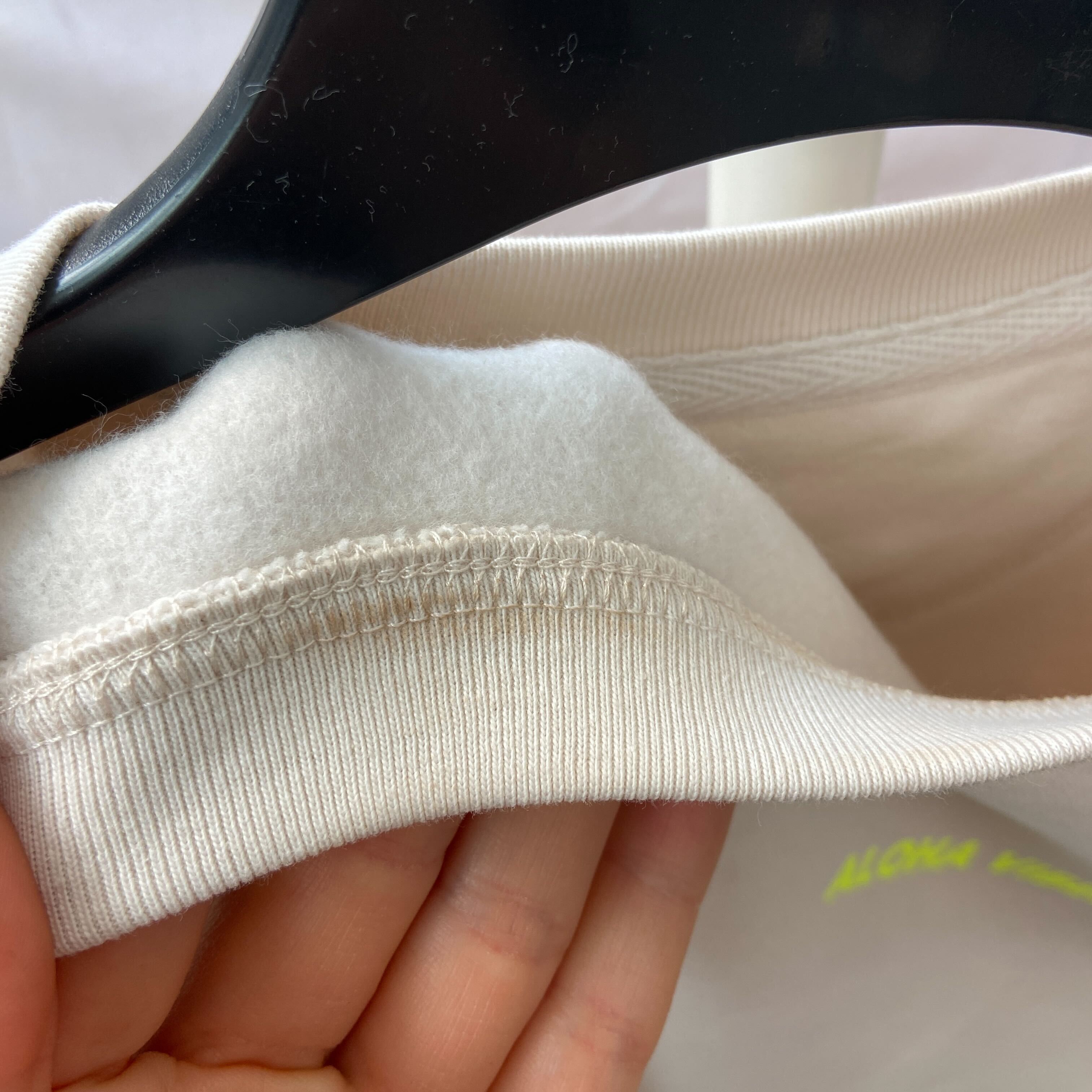 Nahaufnahme einer Hand, die den Kragen eines hellen, mit Fleece gefütterten Sweatshirts hält, wobei die Nähte und der weiche Innenstoff zu sehen sind, während das Kleidungsstück auf einem schwarzen Bügel hängt.