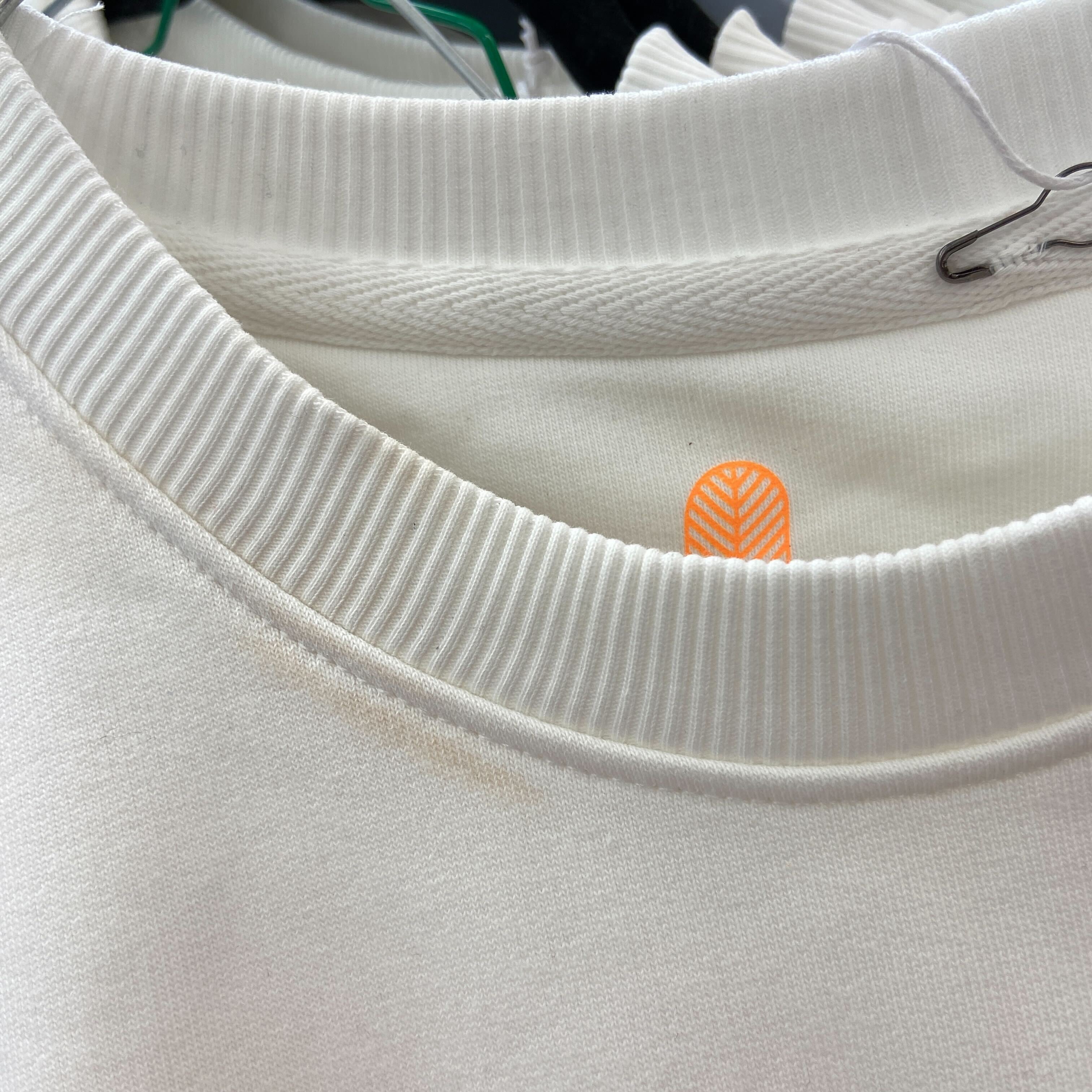 Nahaufnahme des Halsausschnitts eines weißen Pullovers auf einem Kleiderbügel, mit gerippten Details und einem kleinen orangefarbenen Anhänger mit einem Chevron-Muster im Kragen.