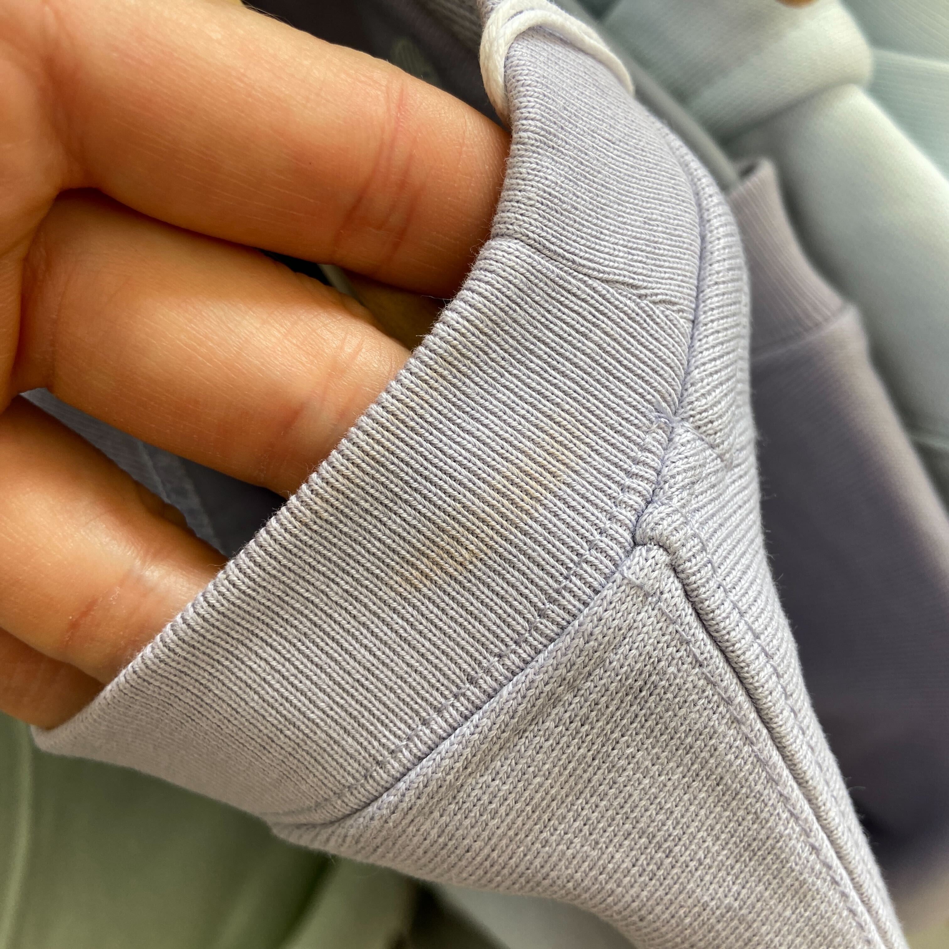 Eine Hand hält die Manschette eines hellvioletten Sweatshirts und zeigt einen schwachen braunen Fleck auf dem Stoff. Die Nahaufnahme konzentriert sich auf die Textur und die Nähte des Bündchens.