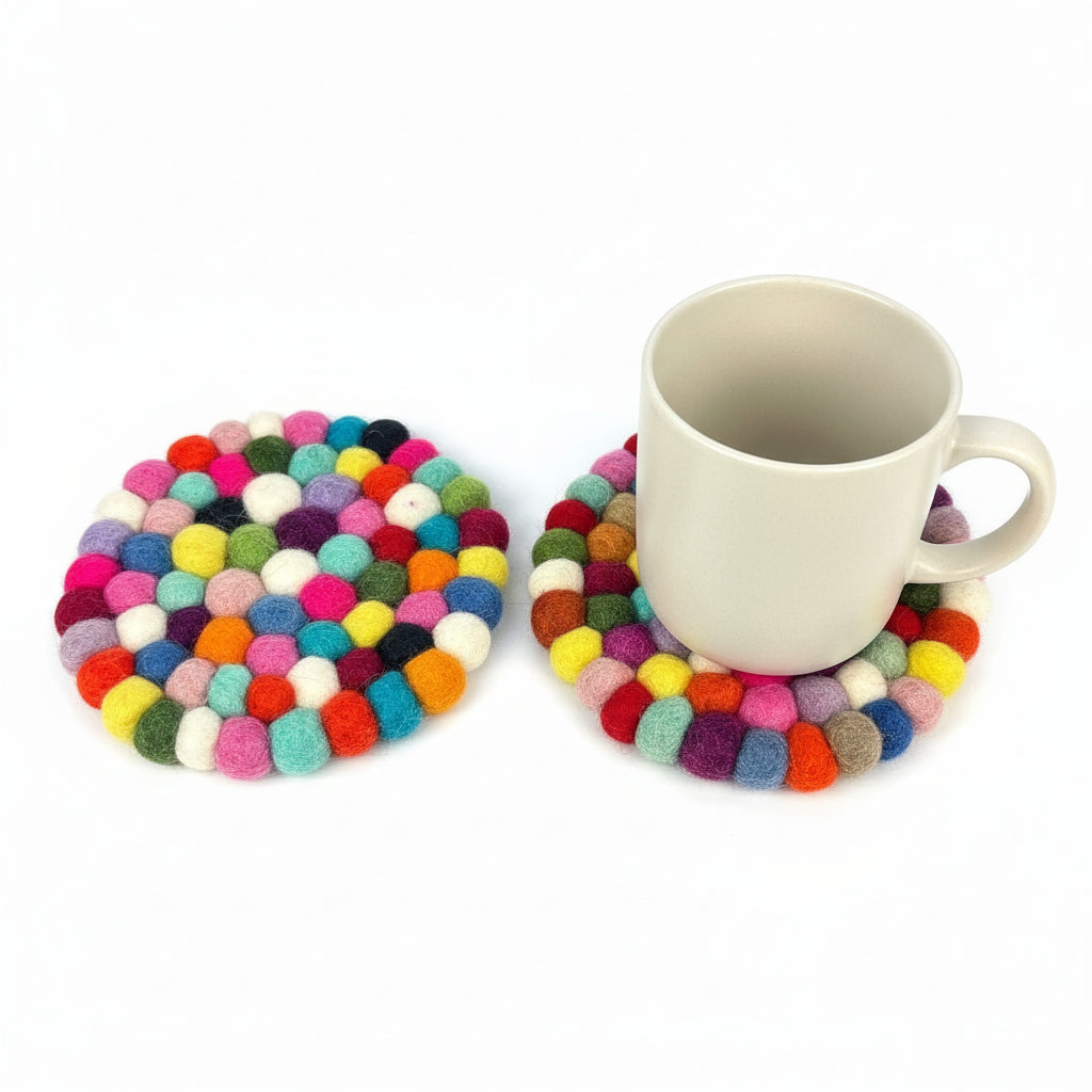 Ein schlichter weißer Becher steht auf einem runden, bunten Untersetzer aus kleinen Filzkugeln, daneben ein weiterer identischer Untersetzer auf weißem Hintergrund.