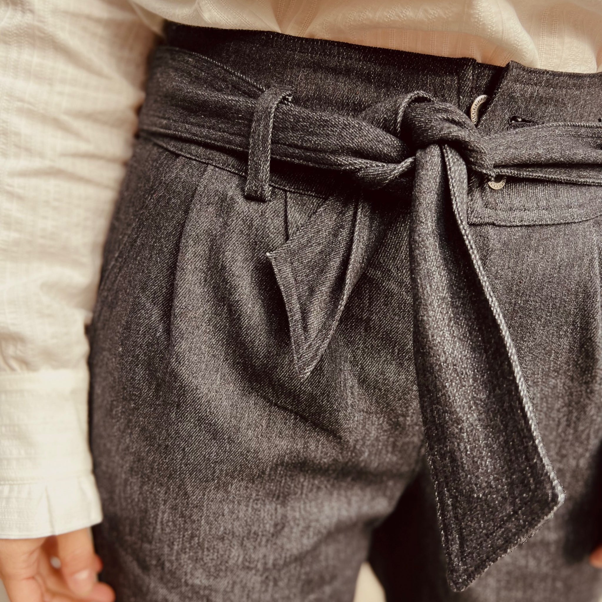 Eine schwarzgraue Hose mit einem Stoffgürtel, der in einer Schleife gebunden ist, wird getragen.