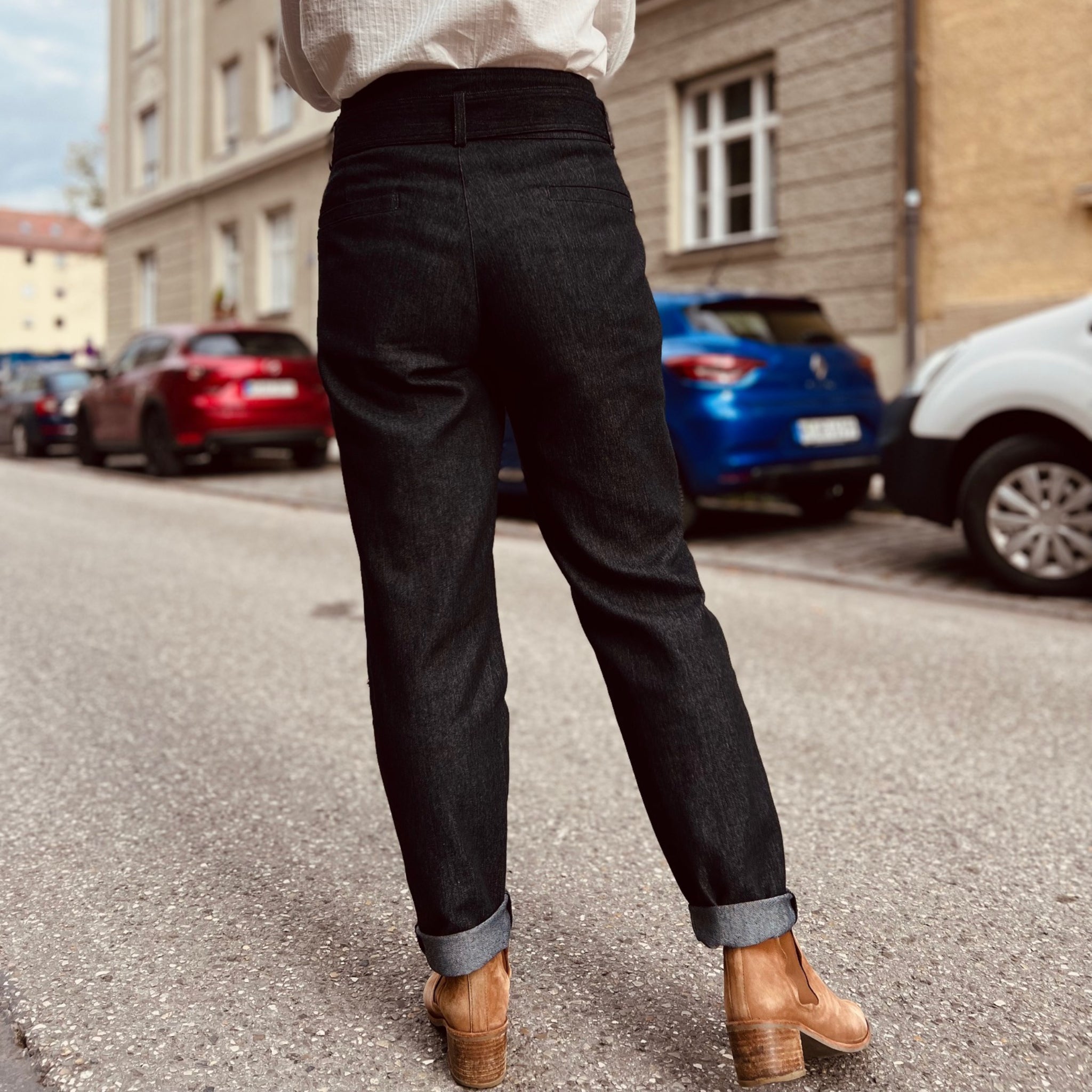 Eine Person in schwarzen Jeans und braunen Stiefeln steht auf einer Straße mit geparkten Autos im Hintergrund.