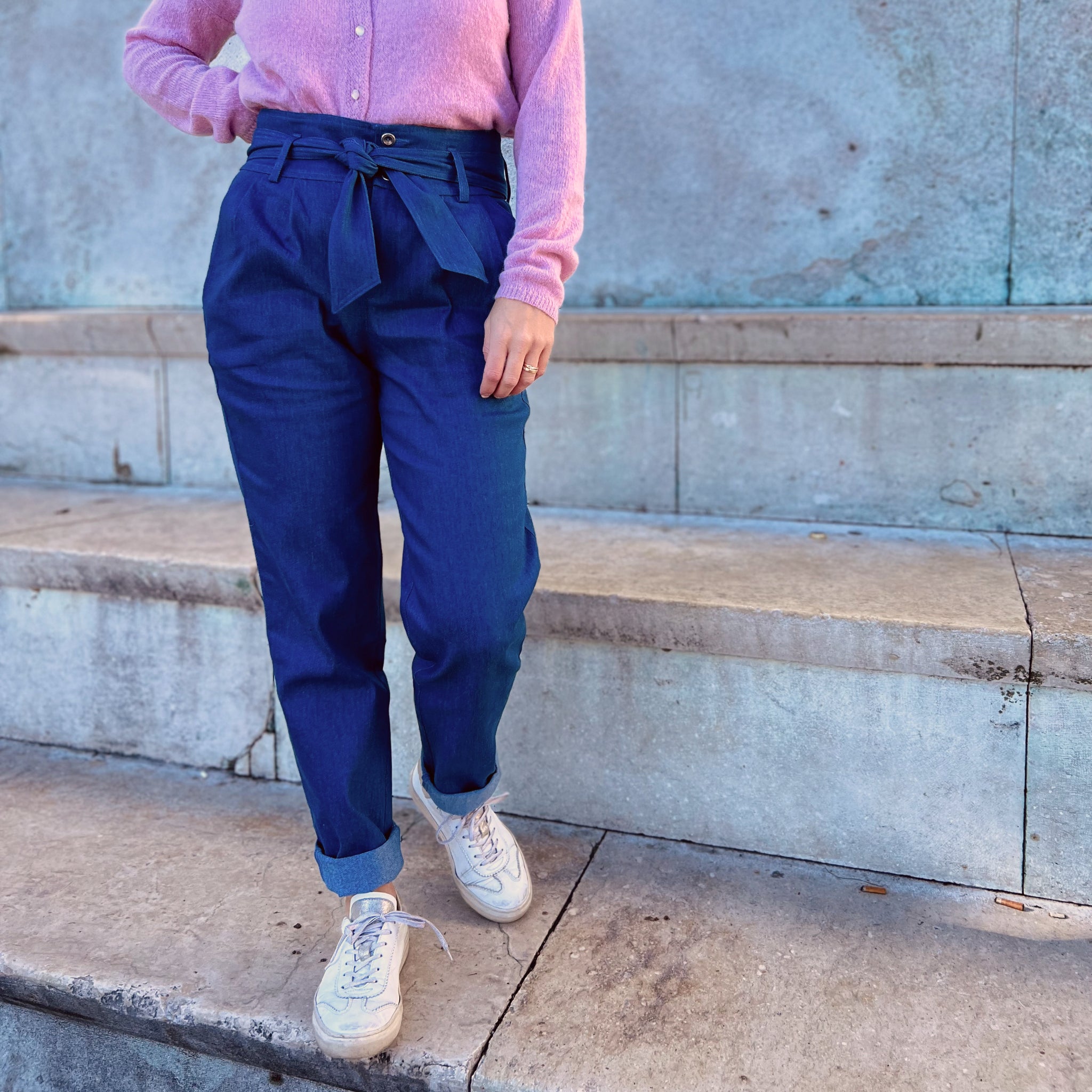 Person in rosa Pullover und blauer Hose mit Gürtel steht auf einer Steintreppe, trägt weiße Sneakers.
