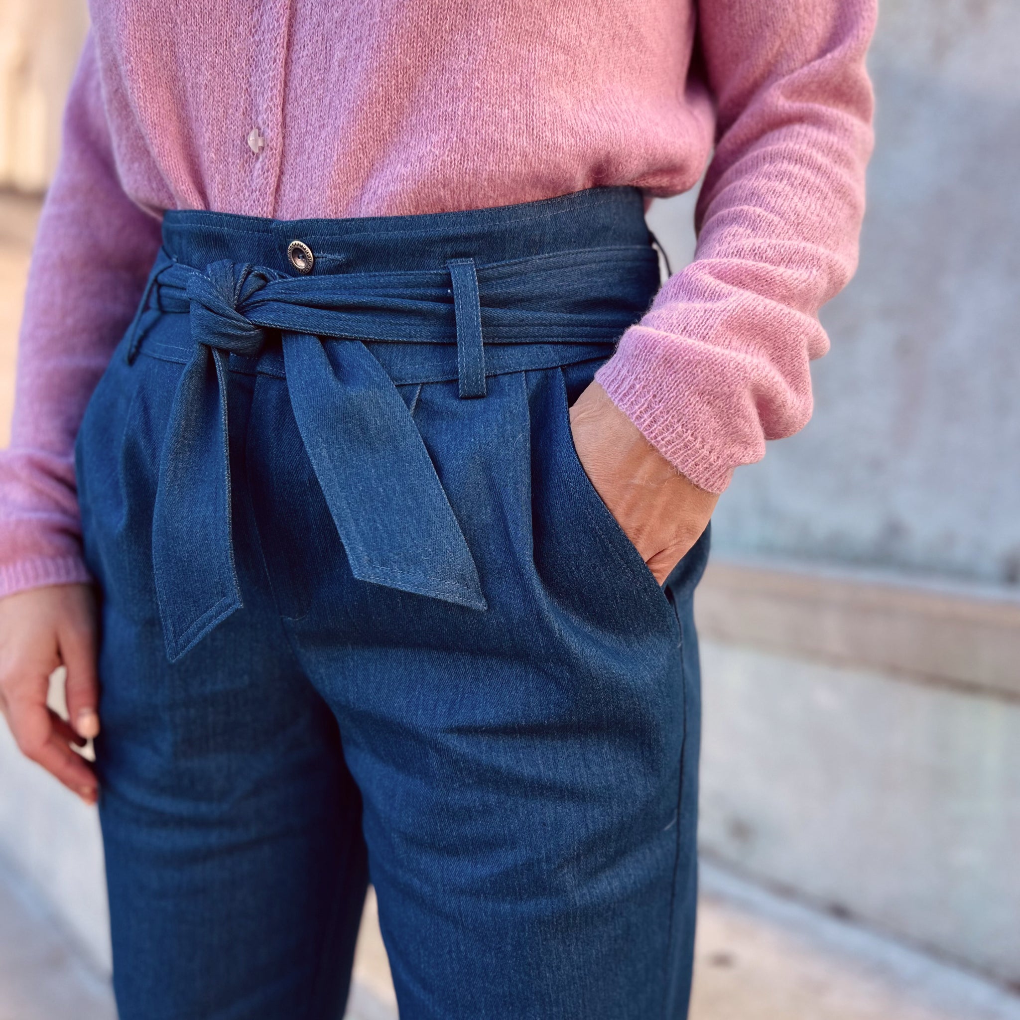 Person trägt eine blaue Hose mit Gürtel sowie einen rosa Pullover, Hand in Hosentasche.