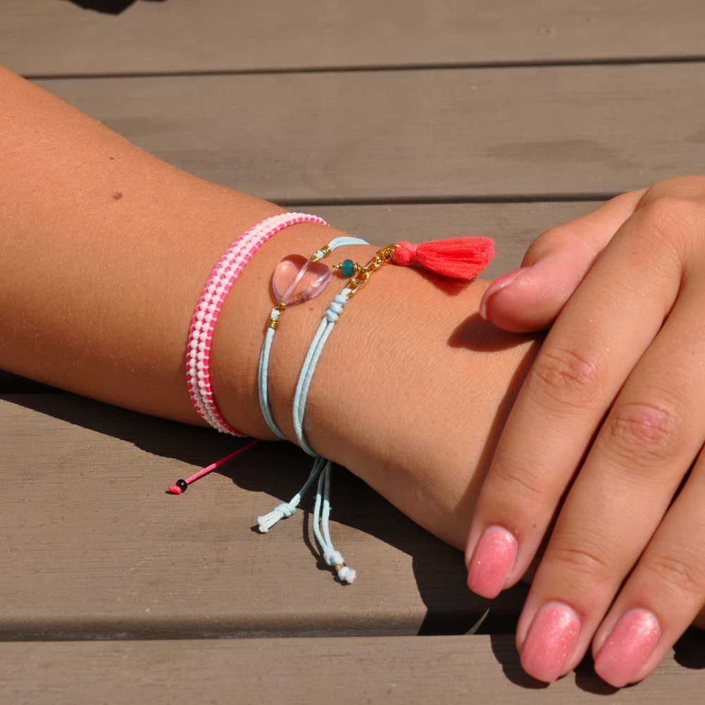 Eine Hand mit rosa lackierten Nägeln ruht auf einer hölzernen Oberfläche und trägt drei bunte Armbänder: ein rosa-weißes Band, eine blaue Kordel mit Perlen und einen roten Quastenanhänger.
