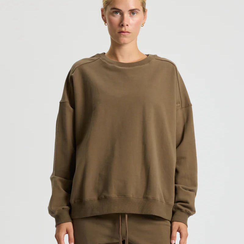 Eine Person mit zurückgebundenem blondem Haar trägt ein locker sitzendes braunes Sweatshirt und eine dazu passende braune Hose und steht vor einem schlichten hellgrauen Hintergrund.