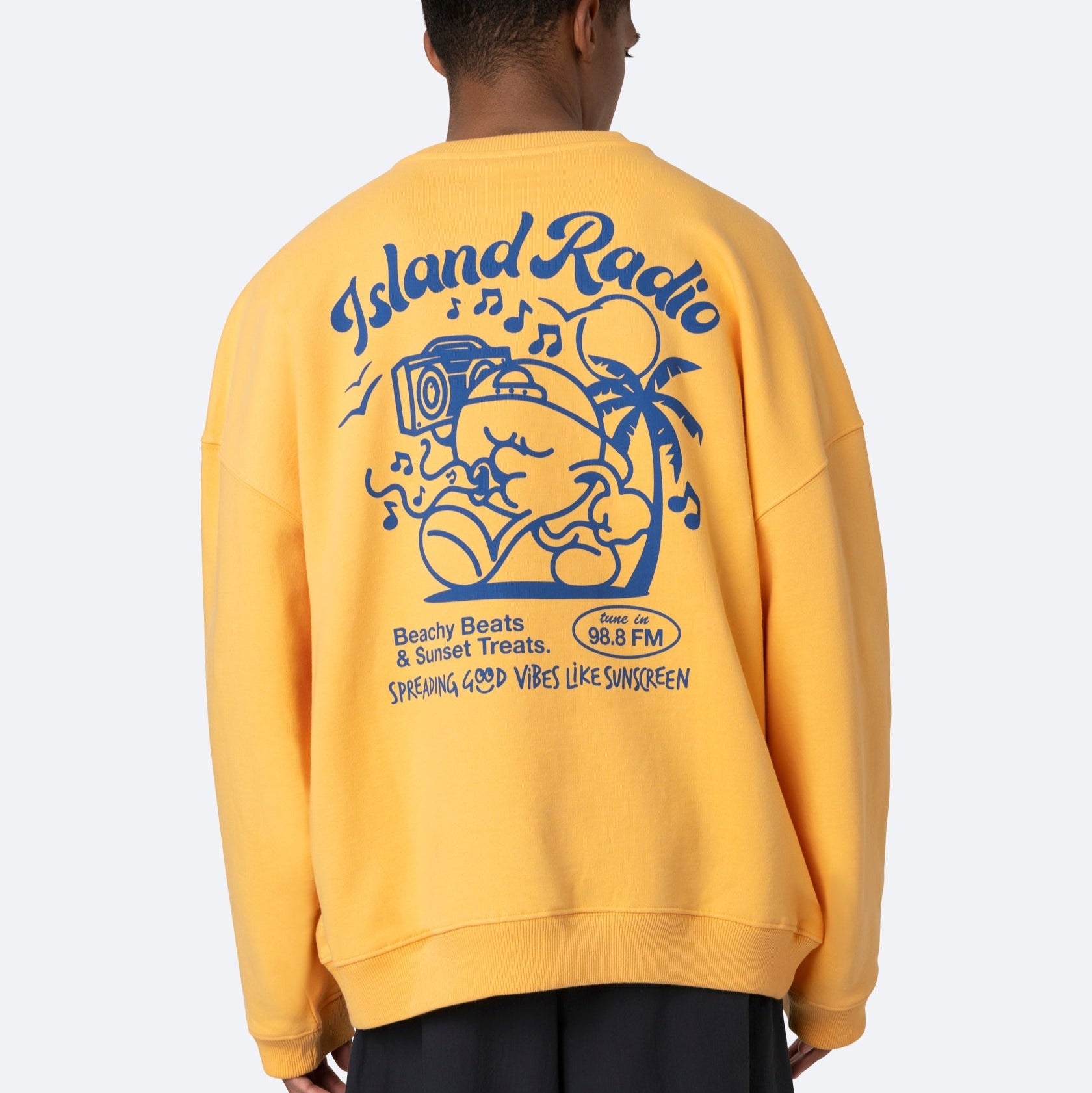 Eine Person, die ein gelbes Sweatshirt mit einer großen blauen Grafik auf dem Rücken trägt, auf der der Schriftzug Island Radio, eine Zeichentrickfigur mit einem Ghettoblaster, Palmen und ein Text über Strandbeats und Sonnenuntergänge zu sehen sind.