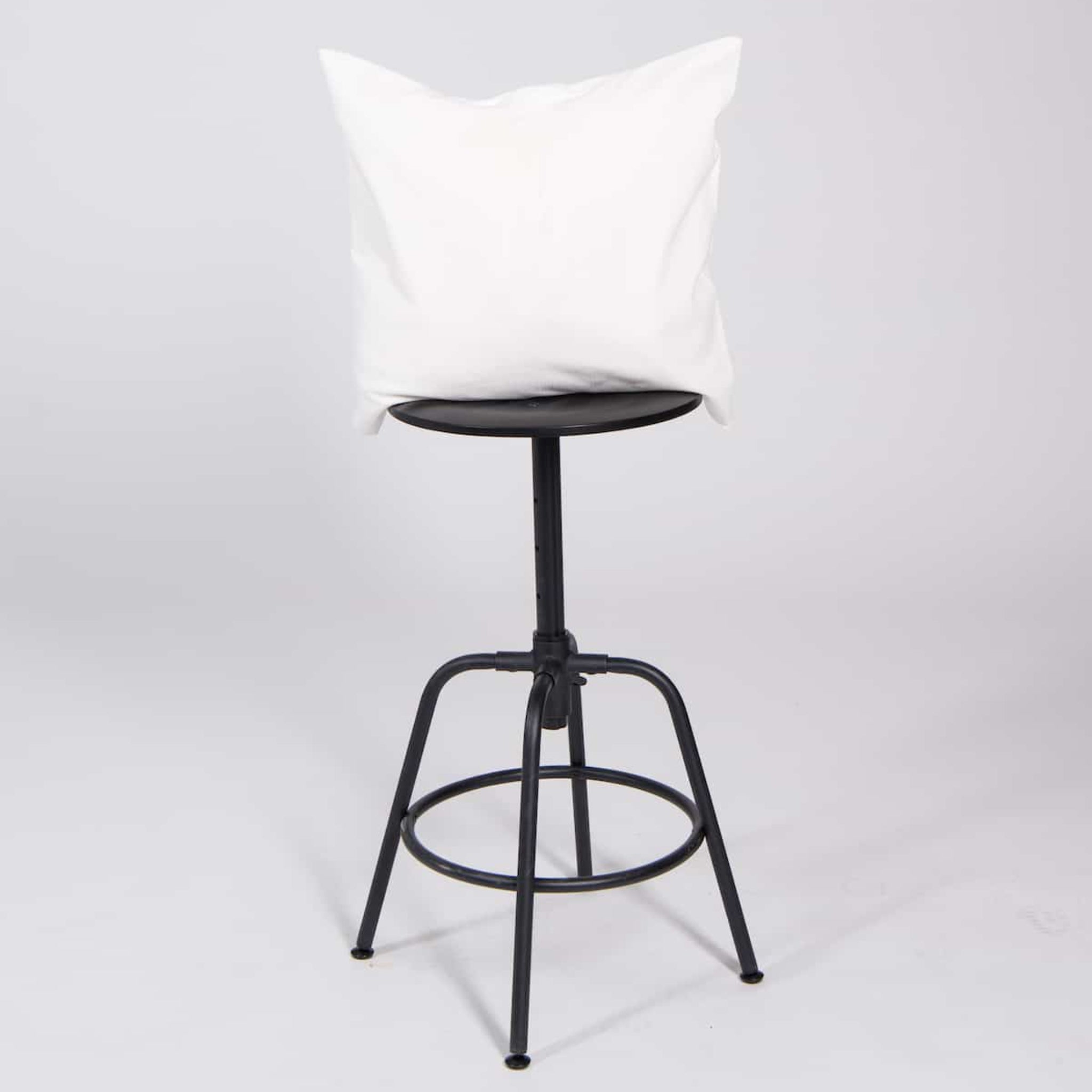 Ein schwarzer Hocker mit rundem Sitz, auf dem ein großes weißes Kissen liegt, vor neutralem Hintergrund.