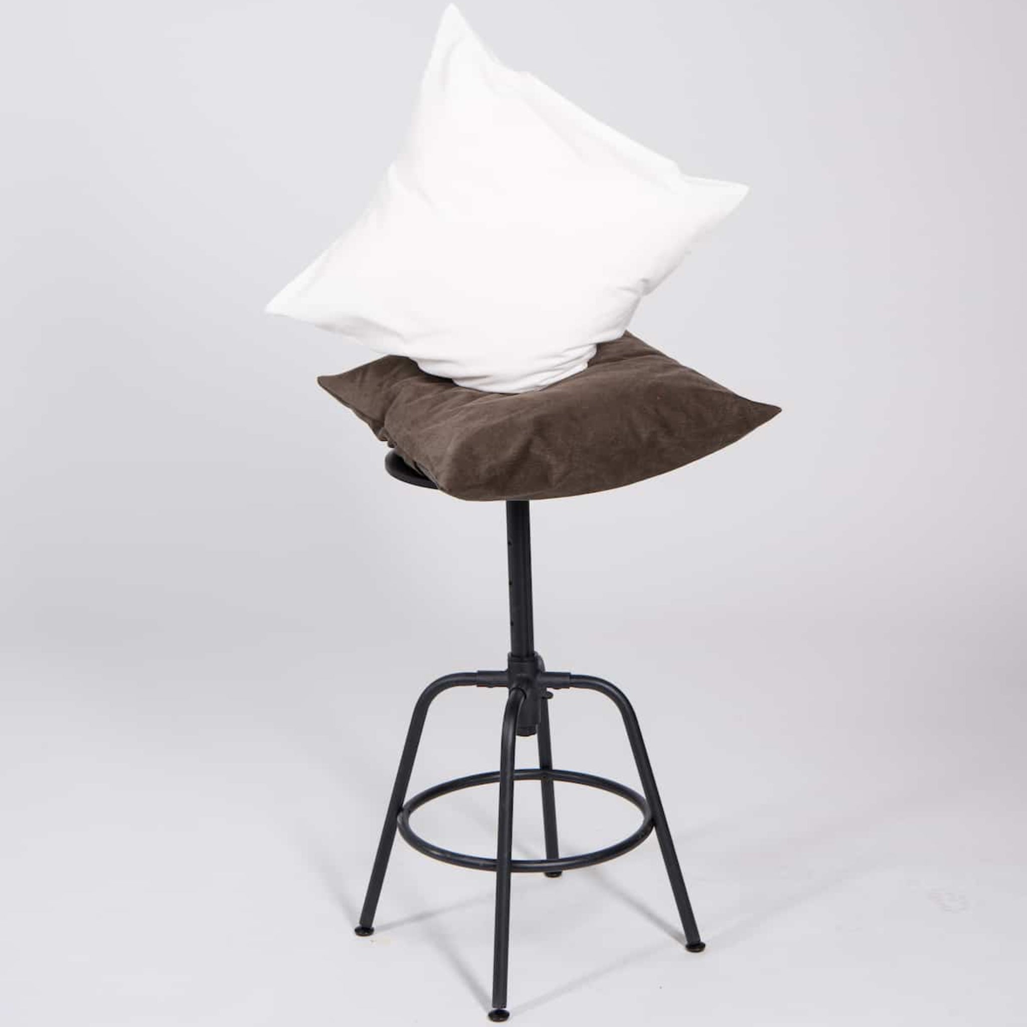 Ein schwarzer Hocker mit einem braunen und einem weißen Kissen darauf, vor einem grauen Hintergrund.