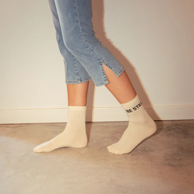 Person trägt blaue Jeans und beige Socken mit der Aufschrift "BE STILL", während sie auf Betonboden geht.