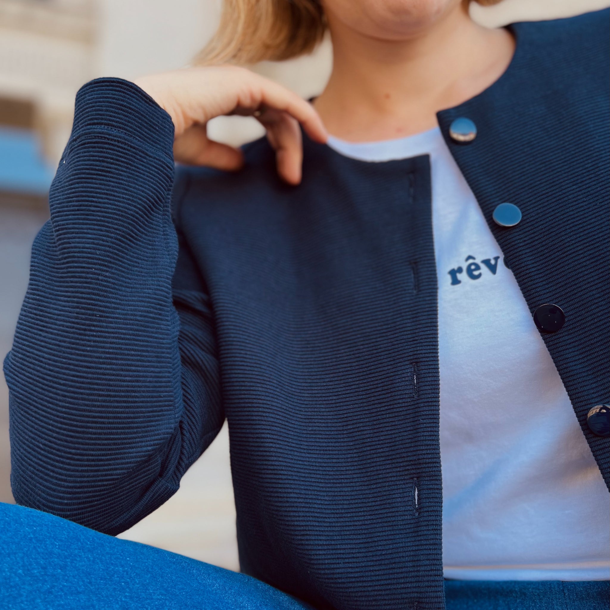 Eine Person trägt eine dunkelblaue Jacke mit silbernen Knöpfen über einem weißen T-Shirt mit dem Wort "rêve".