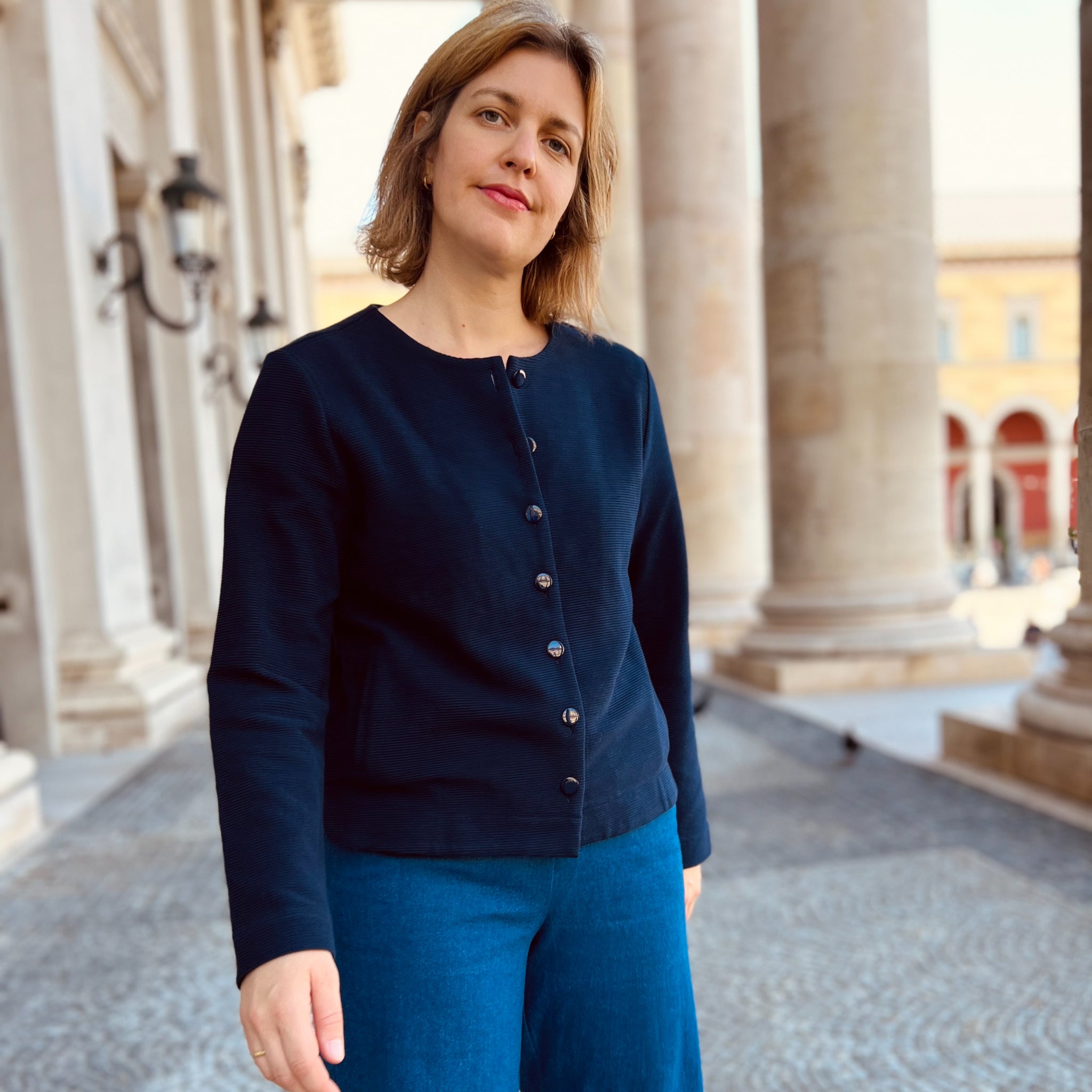Frau in blauer Kleidung steht vor Säulen auf einem gepflasterten Boden, sieht in die Kamera.