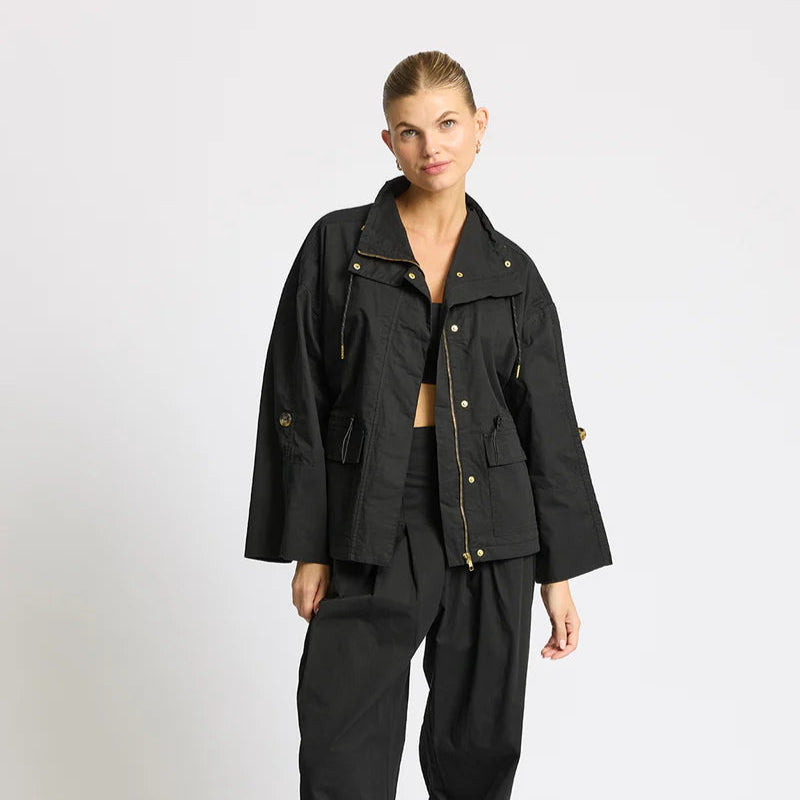 Eine Frau steht vor einem schlichten Hintergrund und trägt eine locker sitzende schwarze Jacke und eine dazu passende schwarze Hose, gepaart mit weiß-braunen Turnschuhen. Sie hat ihr Haar zurückgesteckt und posiert lässig mit einer Hand in ihrer Tasche.