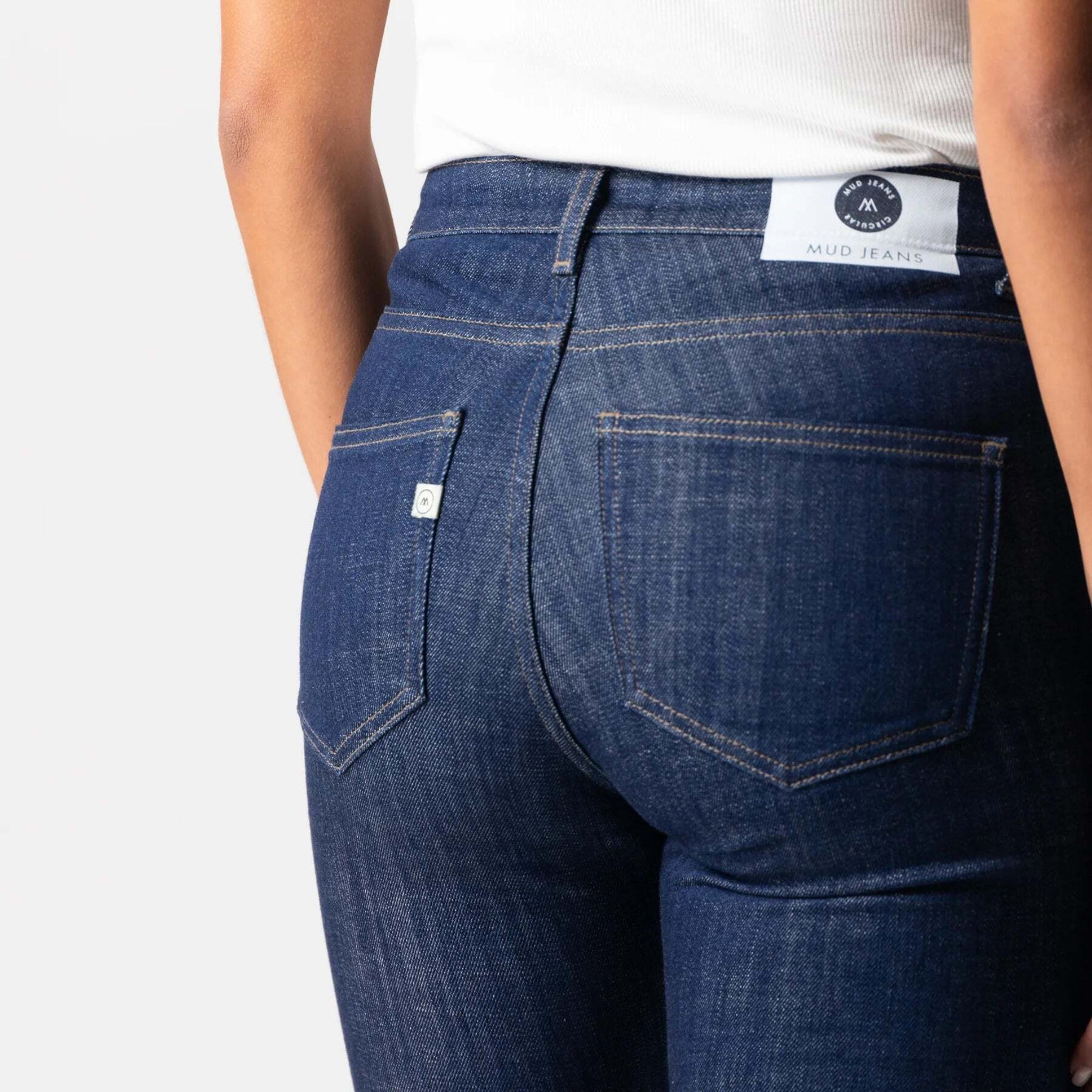 Person in blue jeans from behind, wearing a white top, with a "MUD JEANS" label visible on the waistband.