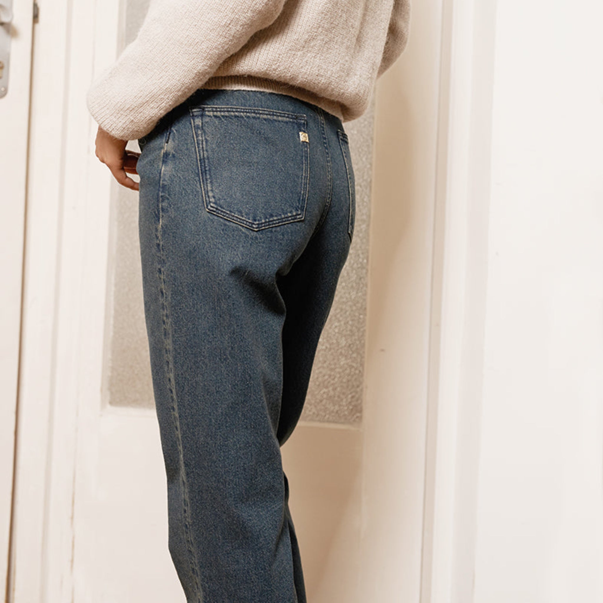 Person trägt blaue Jeans und einen beigen Pullover, fotografiert von hinten in einem Innenraum.