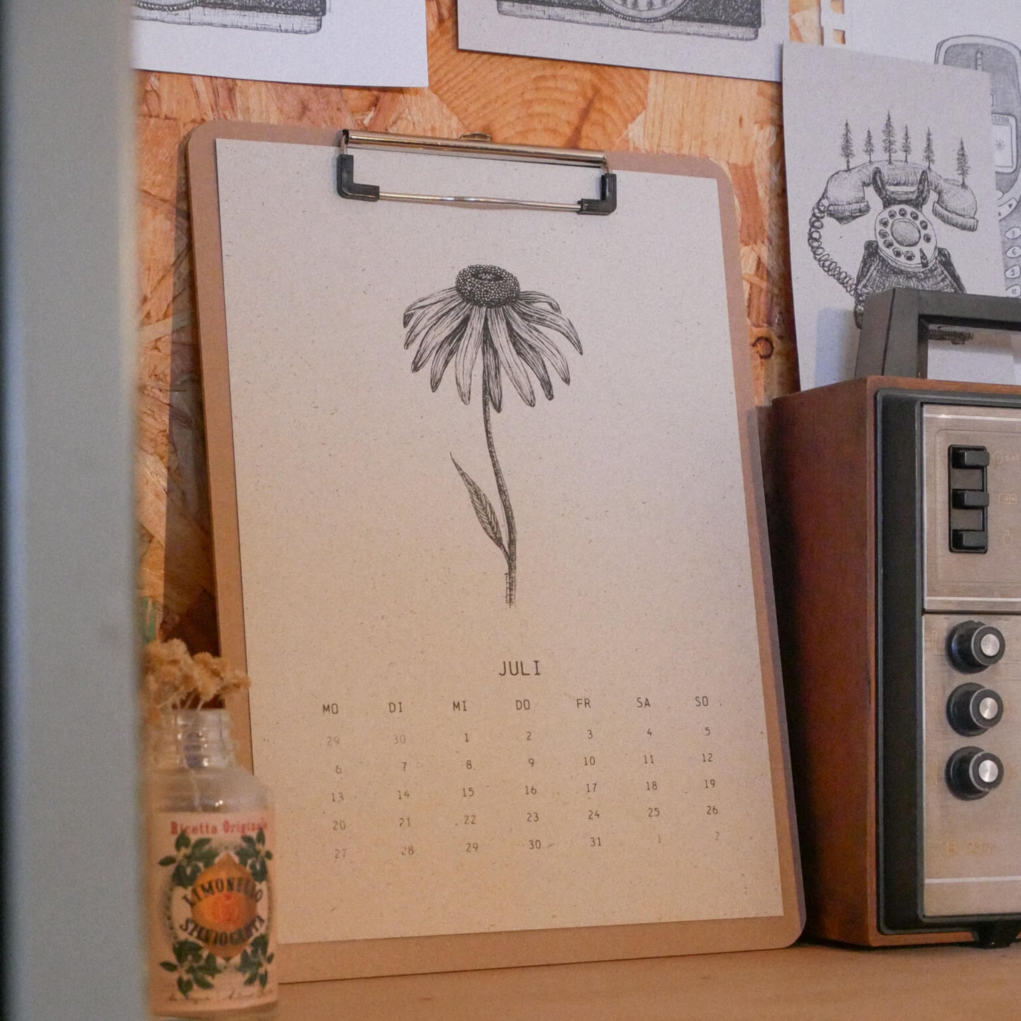 Ein Kalender auf einem Klemmbrett zeigt den Juli mit einer Schwarz-Weiß-Zeichnung einer Blume an. Er steht neben einem alten Radio und einer kleinen Glasflasche auf einer hölzernen Fläche, dahinter sind Skizzen an die Wand gepinnt.