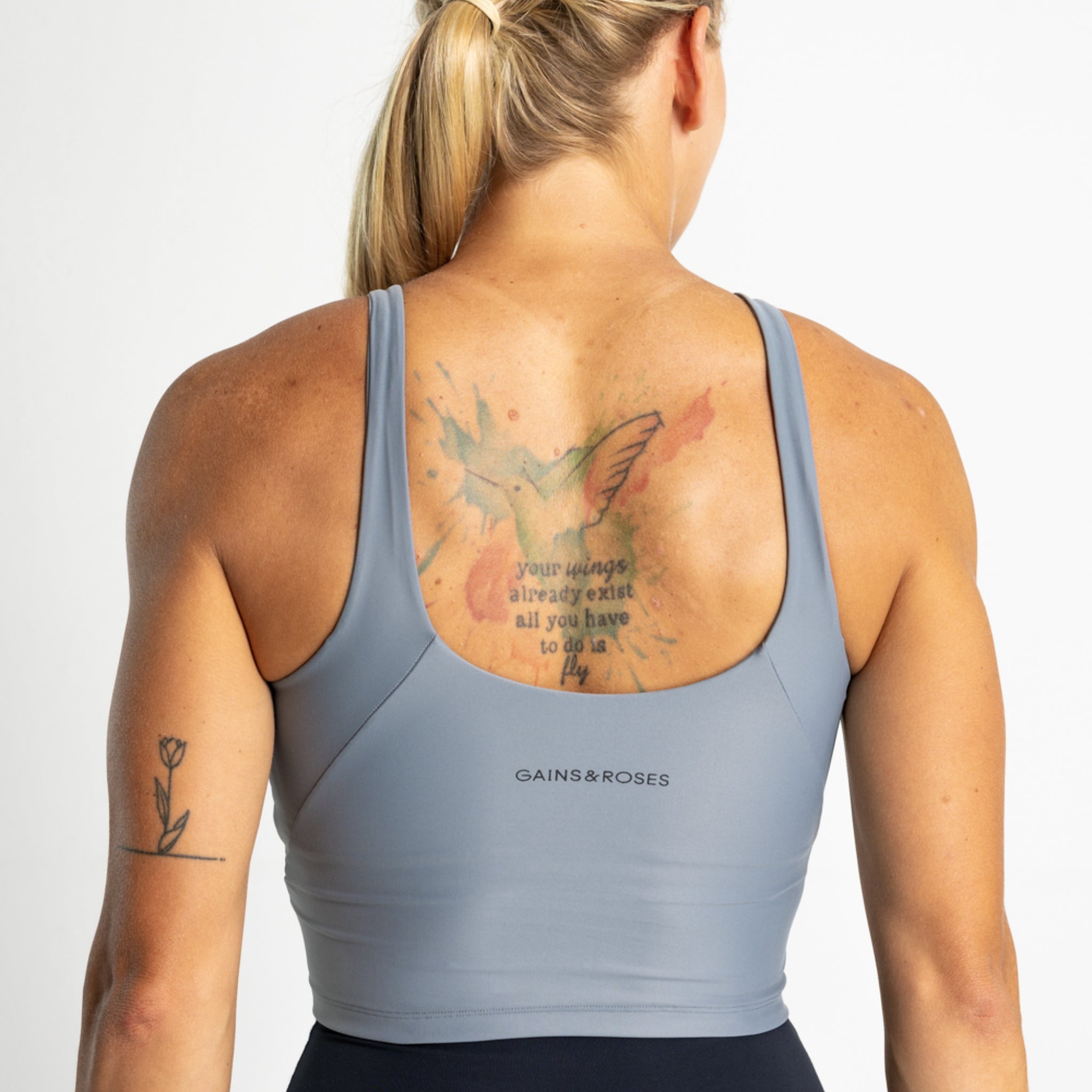 Eine Frau mit einem Tattoo auf dem Rücken trägt ein graues Sporttop und hat eine Tulpe auf dem Arm tätowiert.