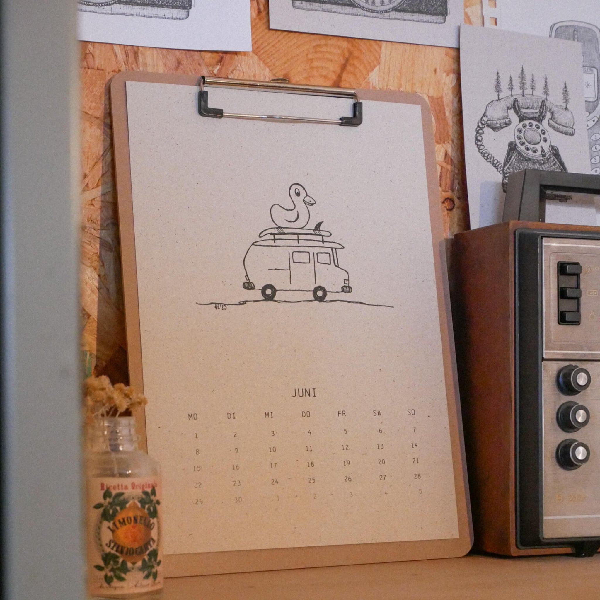 Ein minimalistischer Juni-Kalender auf einem Klemmbrett zeigt eine Strichzeichnung eines Lieferwagens mit einer Ente darauf. Das Klemmbrett liegt auf einem Regal neben einem alten Radio und einer kleinen Flasche mit Trockenblumen. Darüber hängen schwarz-weiße Skizzen.