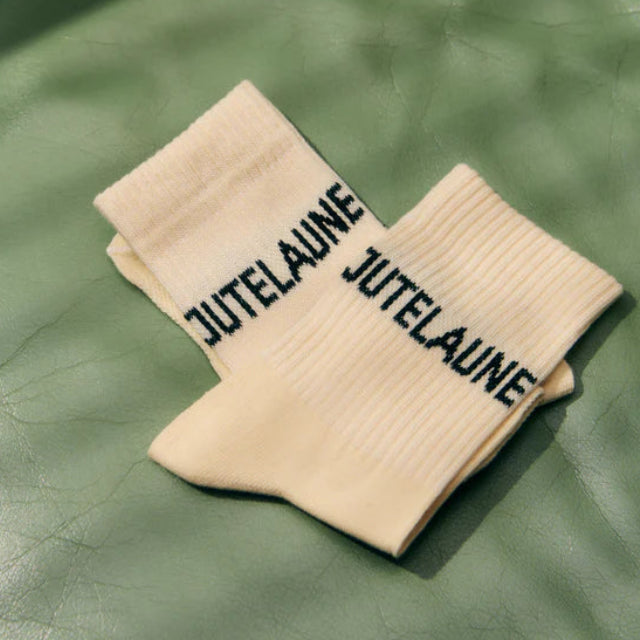 Ein Paar cremefarbene, gerippte Socken mit dem Wort JUTELAUNE in schwarzen Buchstaben auf einer hellgrünen Lederoberfläche.