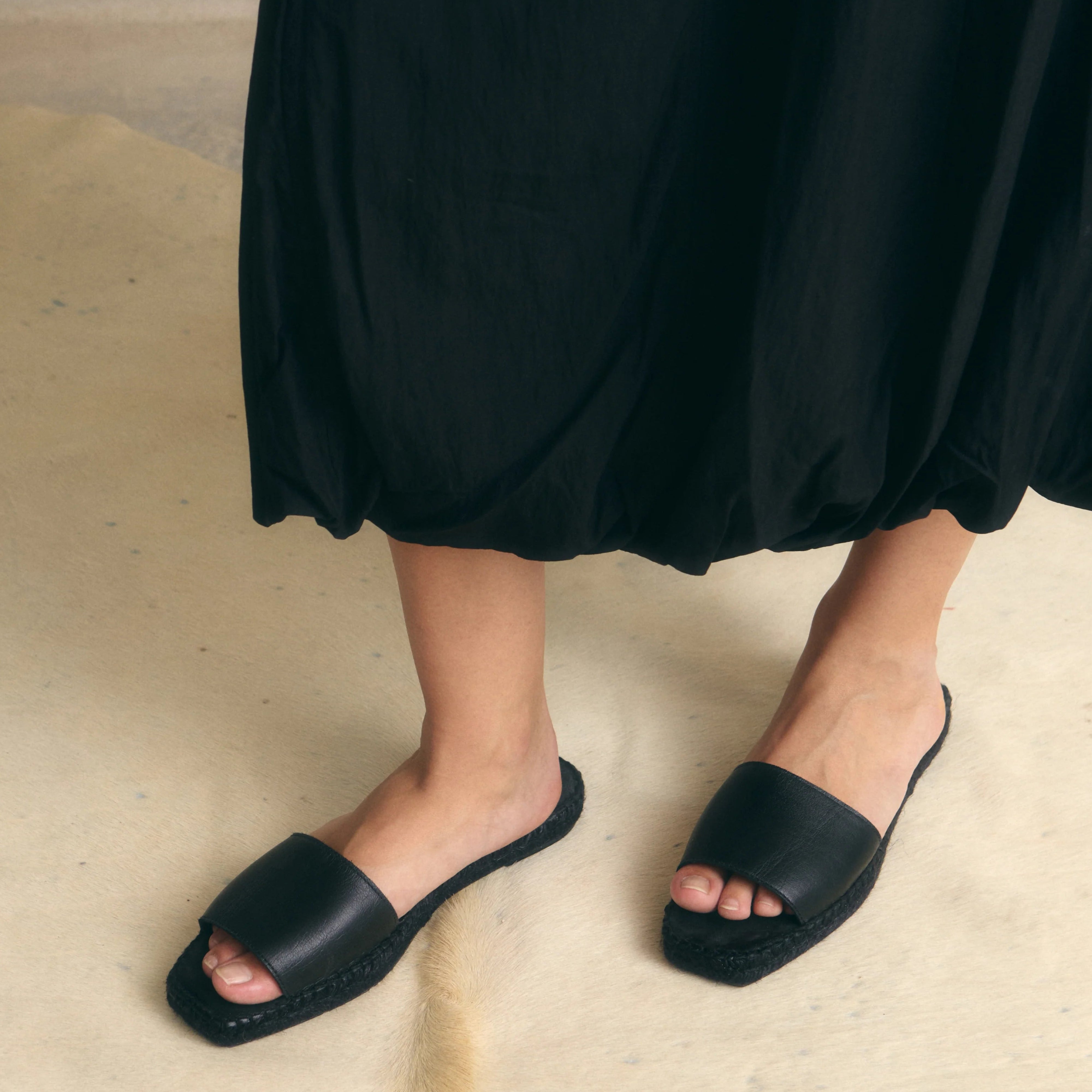 Eine Person, die einen weiten schwarzen Rock und schwarze, offene Sandalen trägt, steht auf einer hellen, strukturierten Fläche. Nur die Unterschenkel und Füße sind sichtbar.