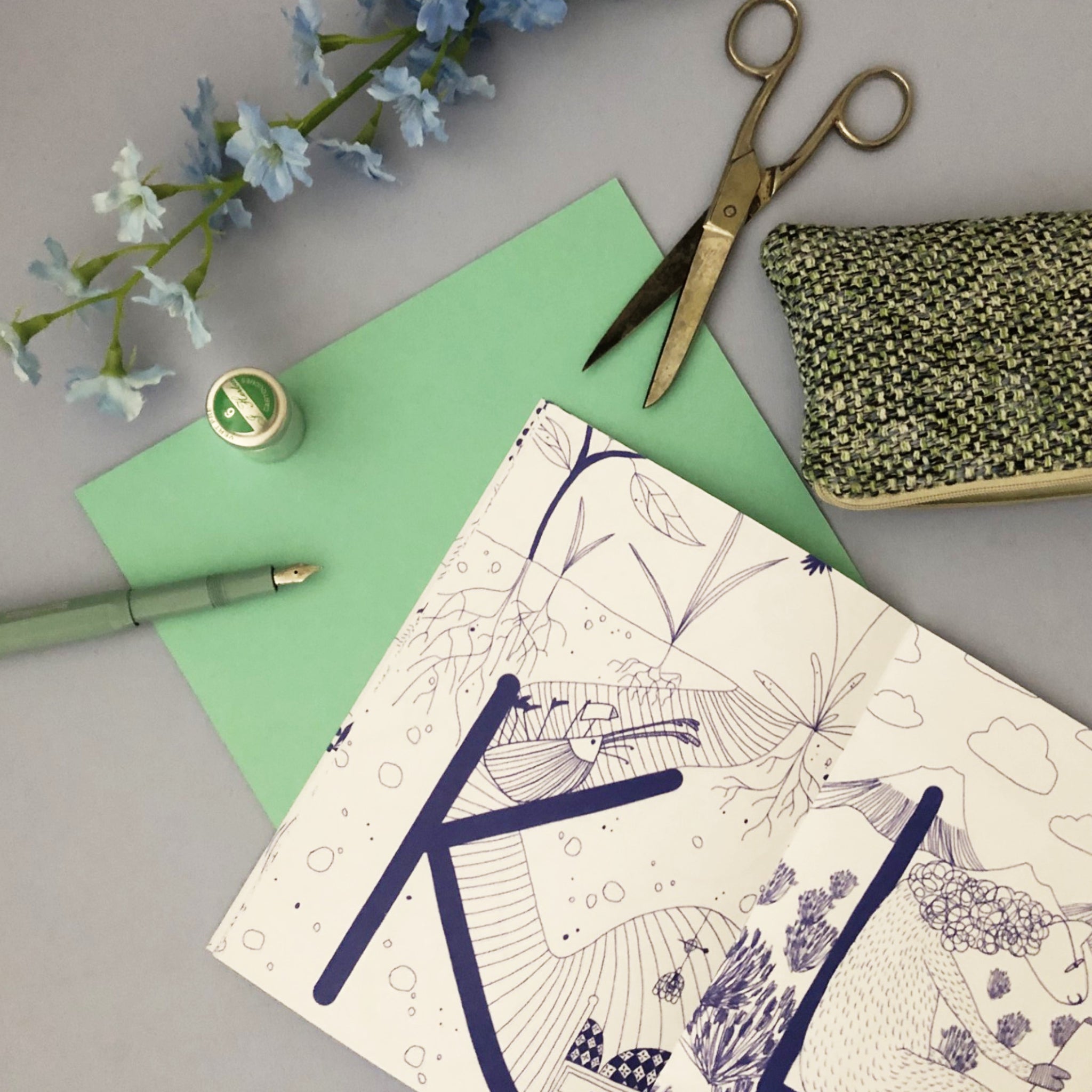 Ein offenes Notizbuch, eine grüne Karte, ein grüner Stift, eine Schere, ein Nähset und blaue Blumen auf einem Tisch.