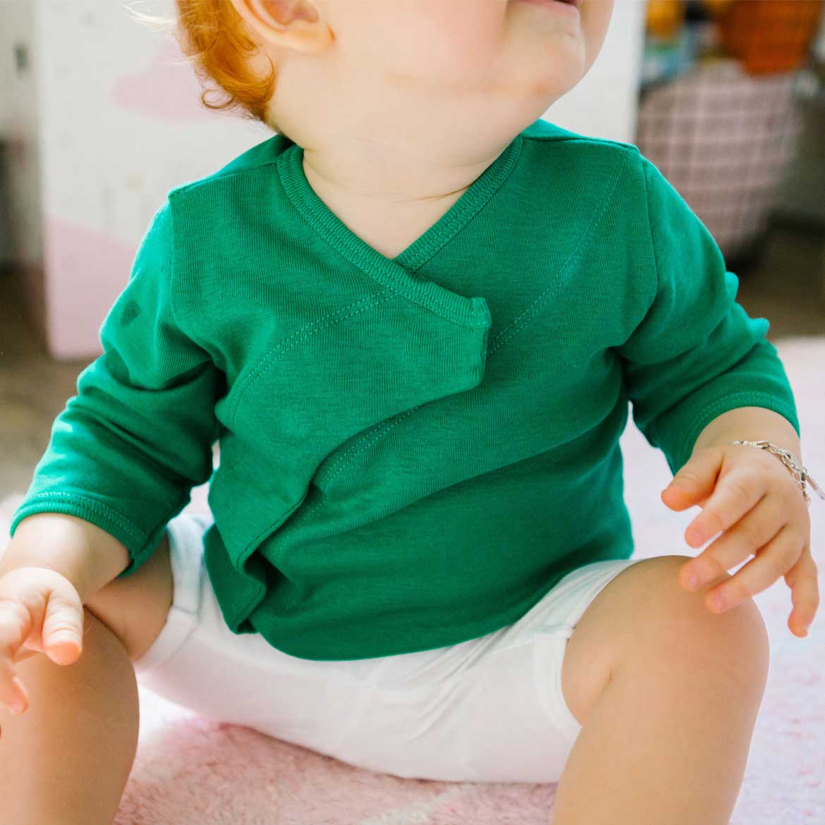 Ein Kleinkind in einem grünen Pullover und weißen Shorts sitzt auf einem Teppich.