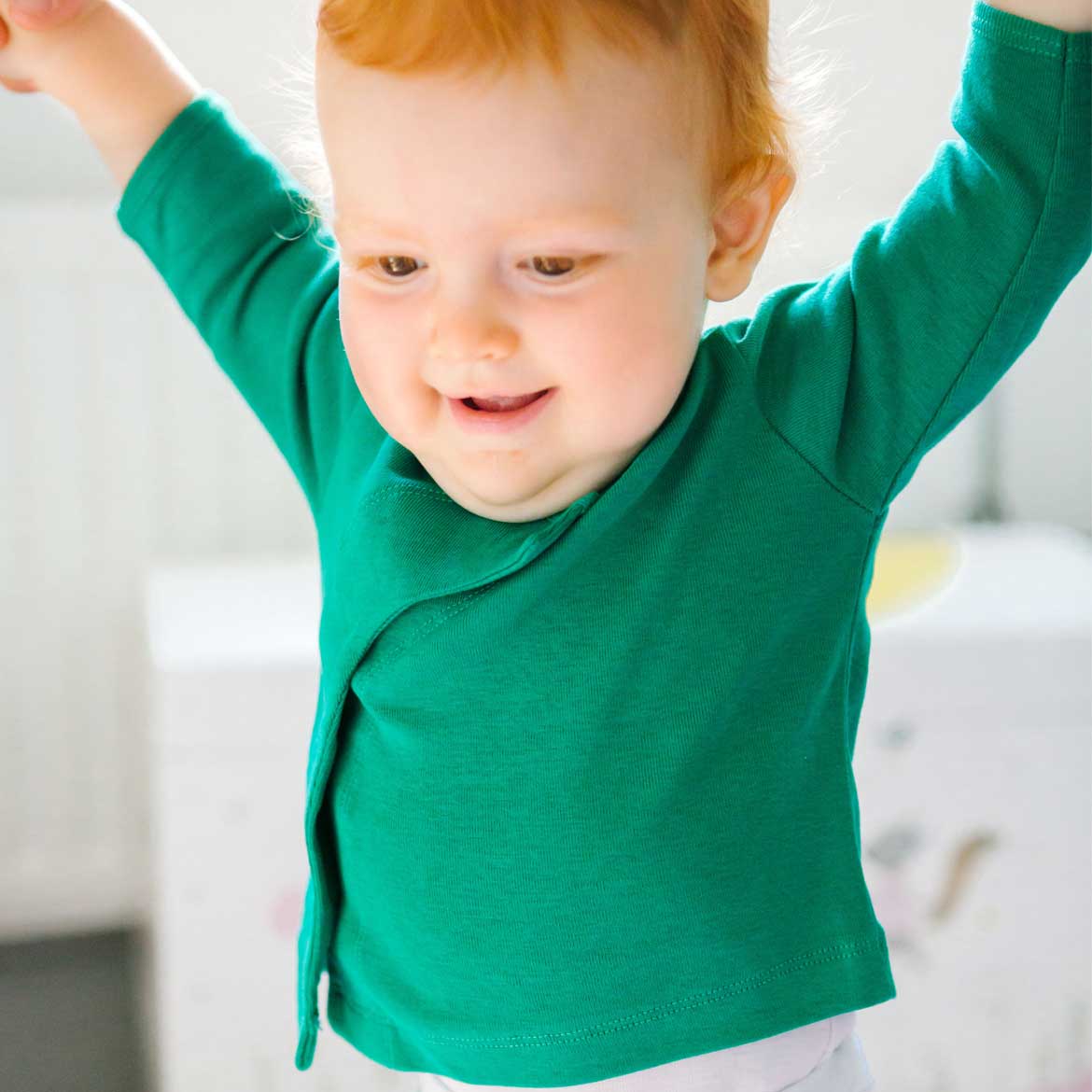Ein lächelndes Baby mit roten Haaren trägt ein grünes Oberteil und hält die Arme in die Luft.