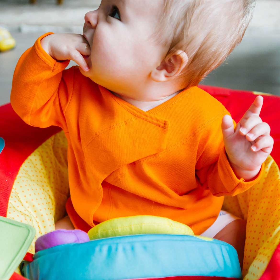 Ein Baby in einem orangefarbenen Pullover sitzt in einem bunten Sitzring und steckt eine Hand in den Mund.