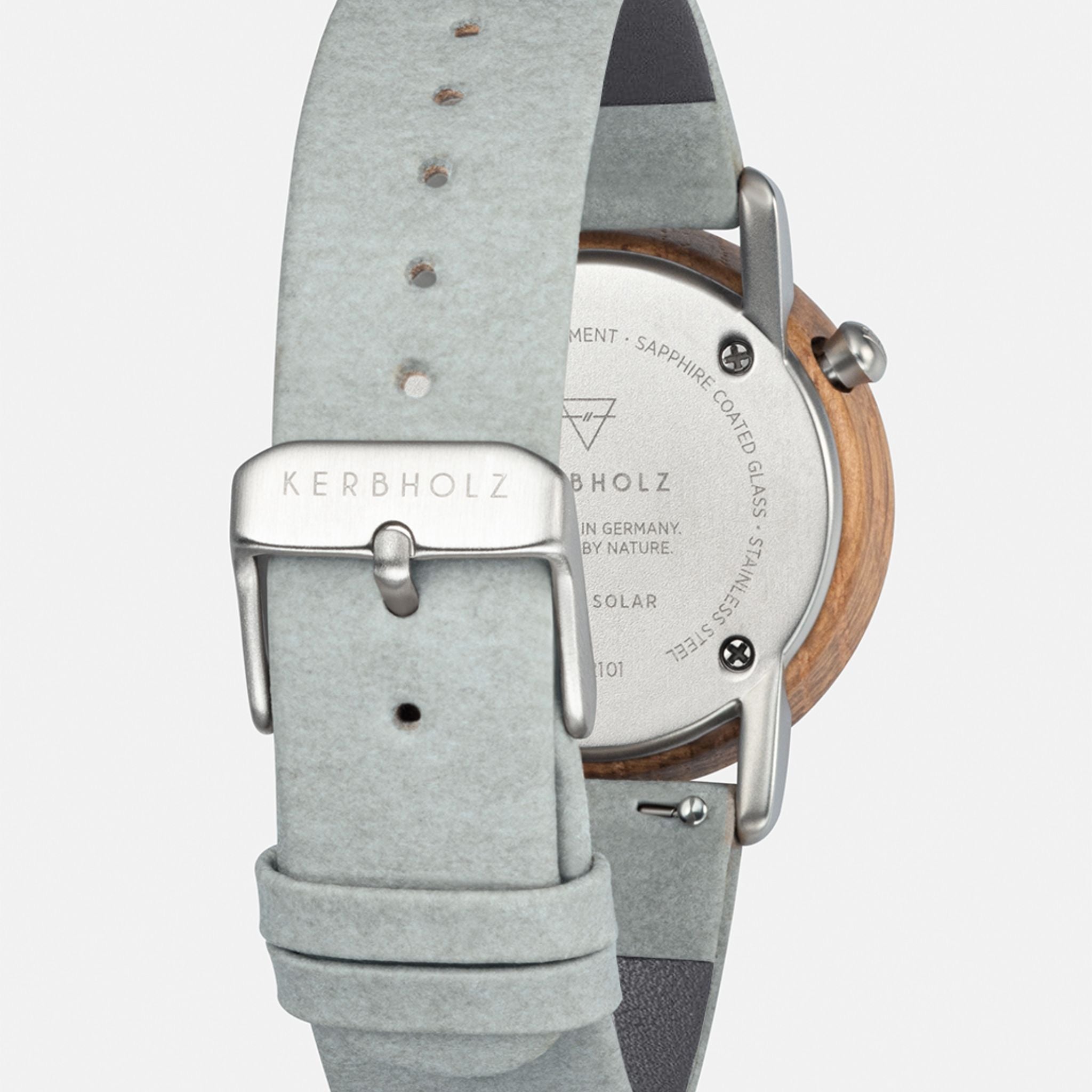 Rückseite einer Kerbholz-Uhr mit grauem Lederarmband und Metallschließe, Text auf der Uhr erkennbar.
