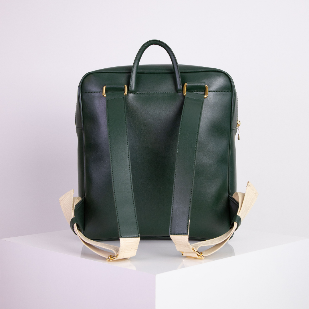 Grüner Lederrucksack mit beigen Trägern und goldenem Zubehör auf einem weißen Podest.